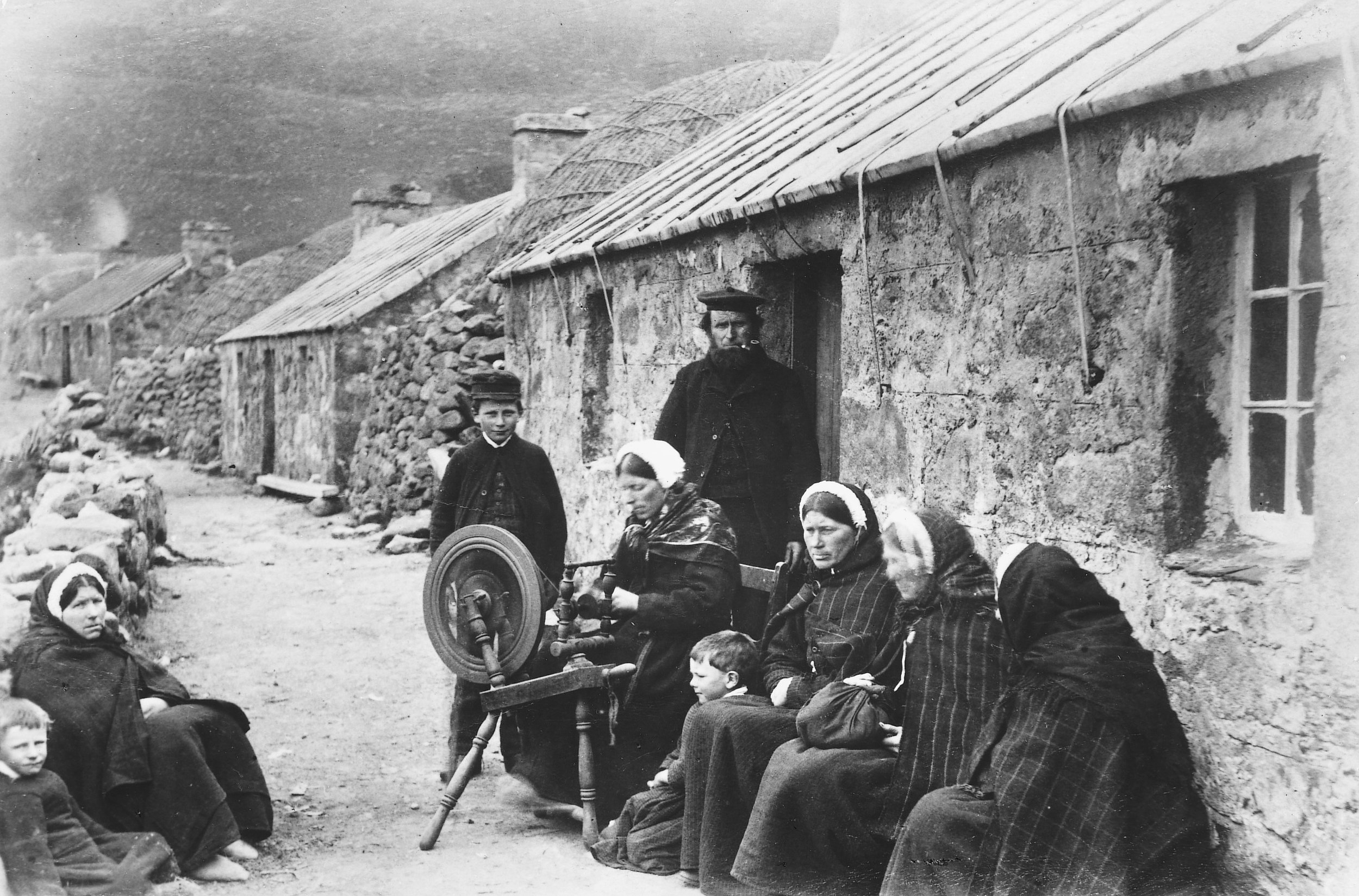 Black and white photo of a group of people sitting on a dirt street outside stone houses in a rural village. A woman is operating a spinning wheel. Several children and women are seated nearby, some wearing headscarves, and a man stands in the doorway of one of the houses.