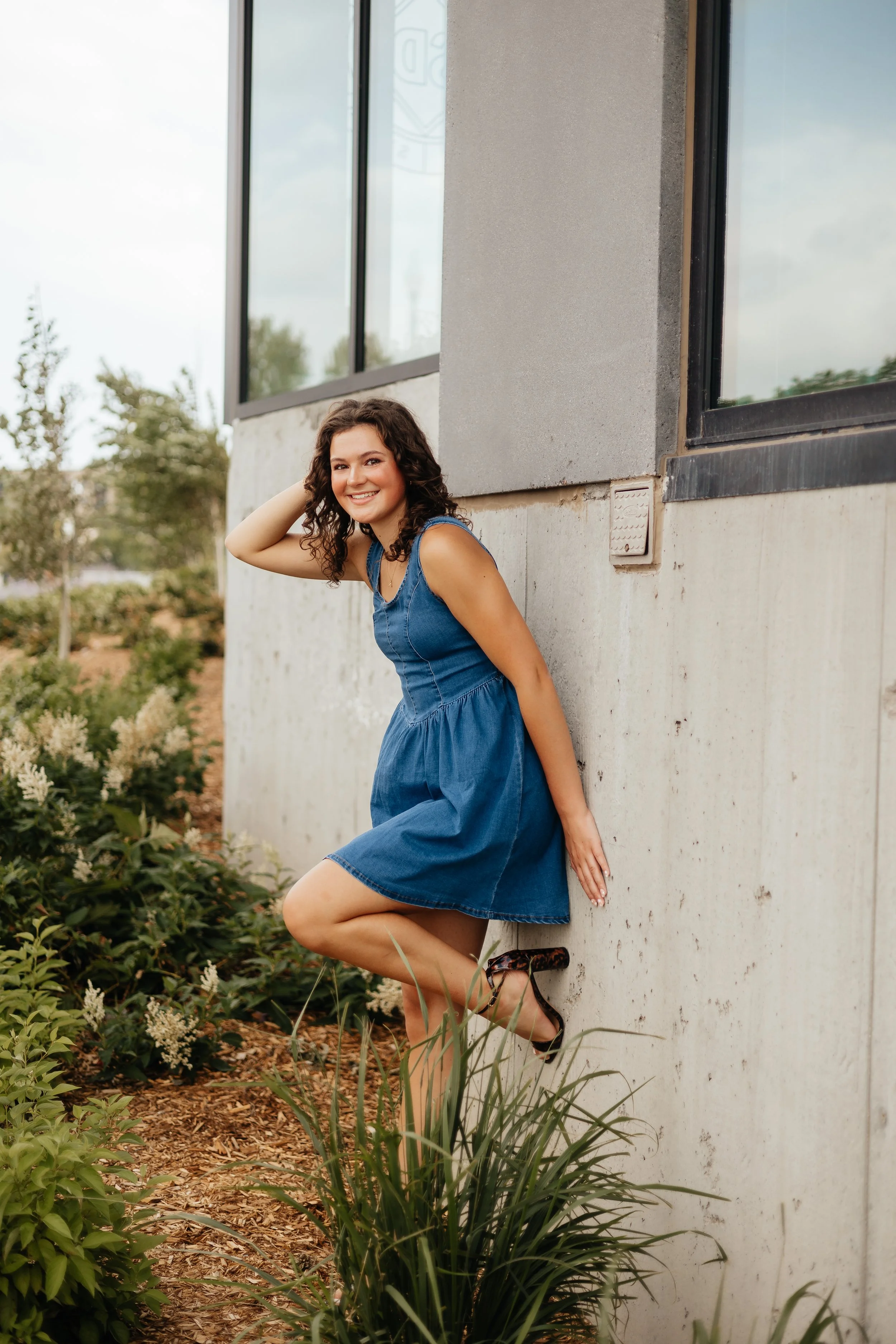 A young woman with curly brown hair smiling, wearing a blue dress, leaning against a concrete wall outdoors with greenery around.
