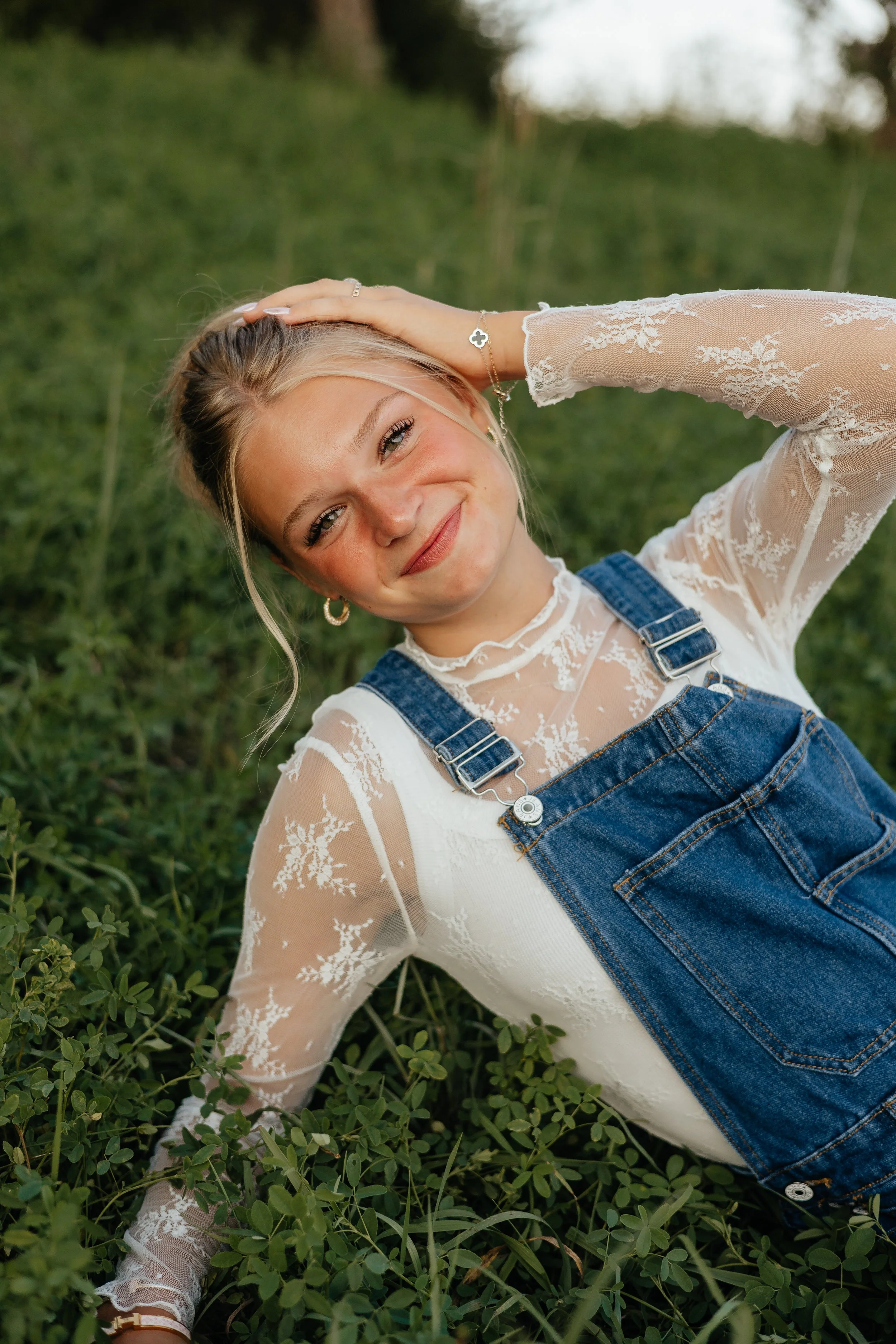 Young woman lying on grass in a field, smiling, with her head resting on one hand, wearing a white lace top and denim overalls.