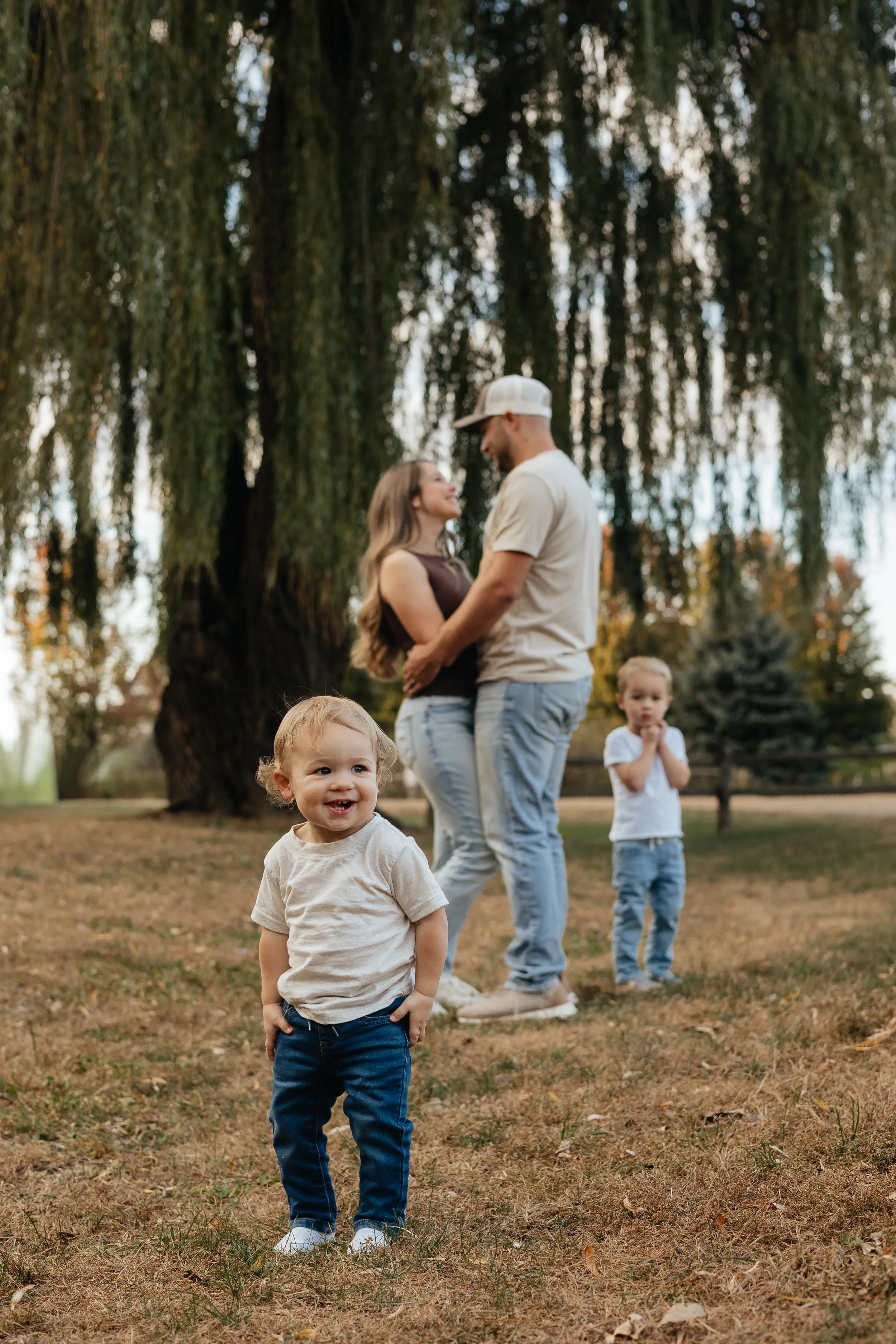 A happy toddler standing in the foreground outdoors with a grassy ground, wearing a beige T-shirt and dark jeans. In the background, a couple with two children, a large tree, and a cloudy sky, appear to be enjoying a family outing at a park or backya