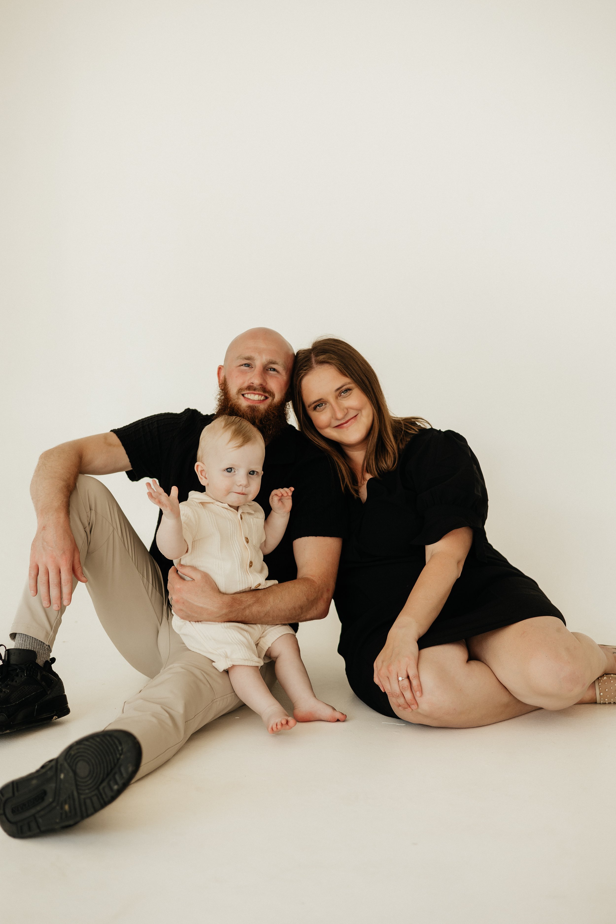 A happy family of three, a man, a woman, and a young child, sitting on the floor against a plain white background. The man has a beard and is smiling, wearing a black shirt and beige pants. The woman has long brown hair, smiling, dressed in black. Th