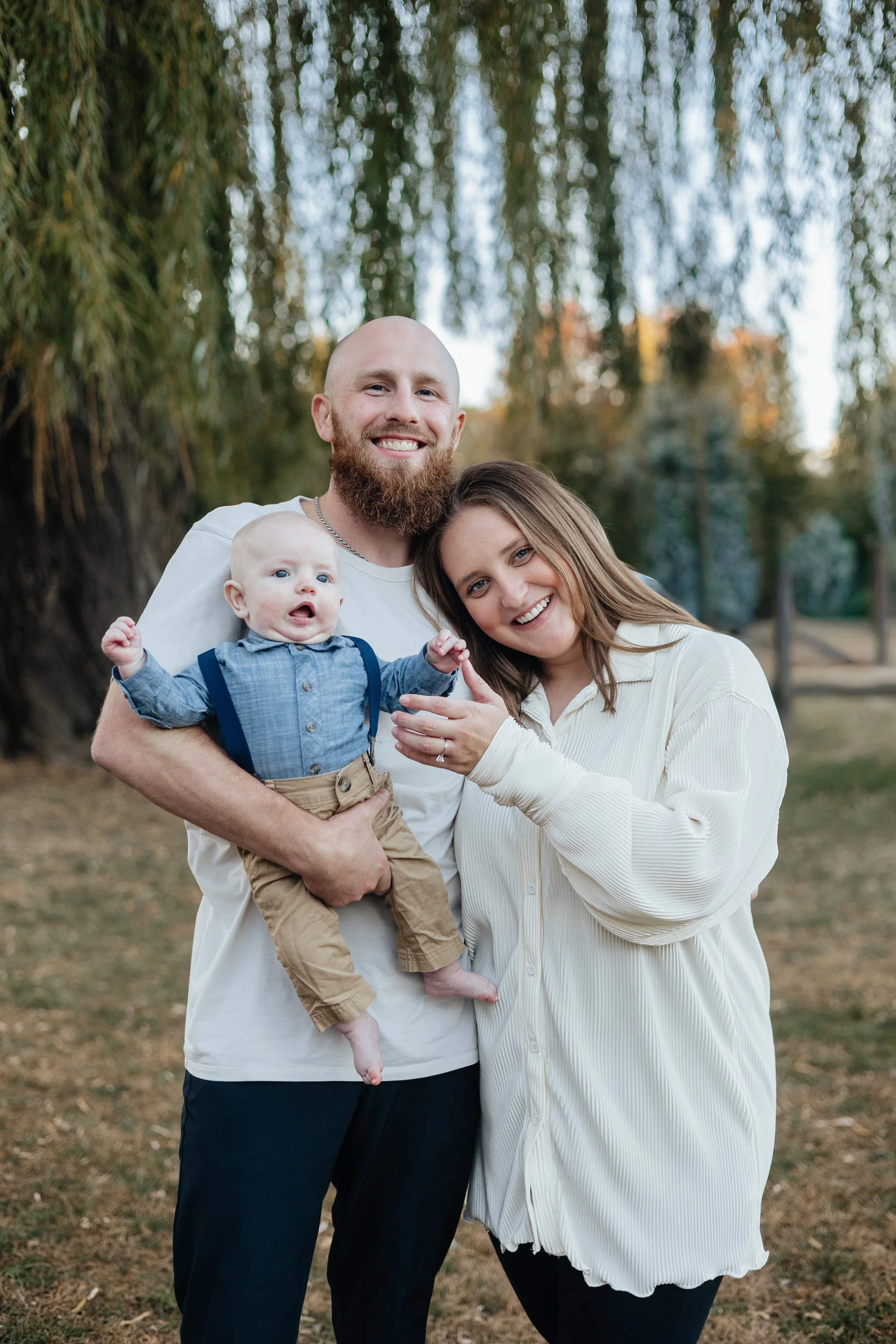 A happy family of three outdoors in a park with tall trees in the background. The father, with a shaved head and beard, is holding their baby boy who has light hair and blue eyes. The mother has long brown hair and is smiling, leaning her head on the