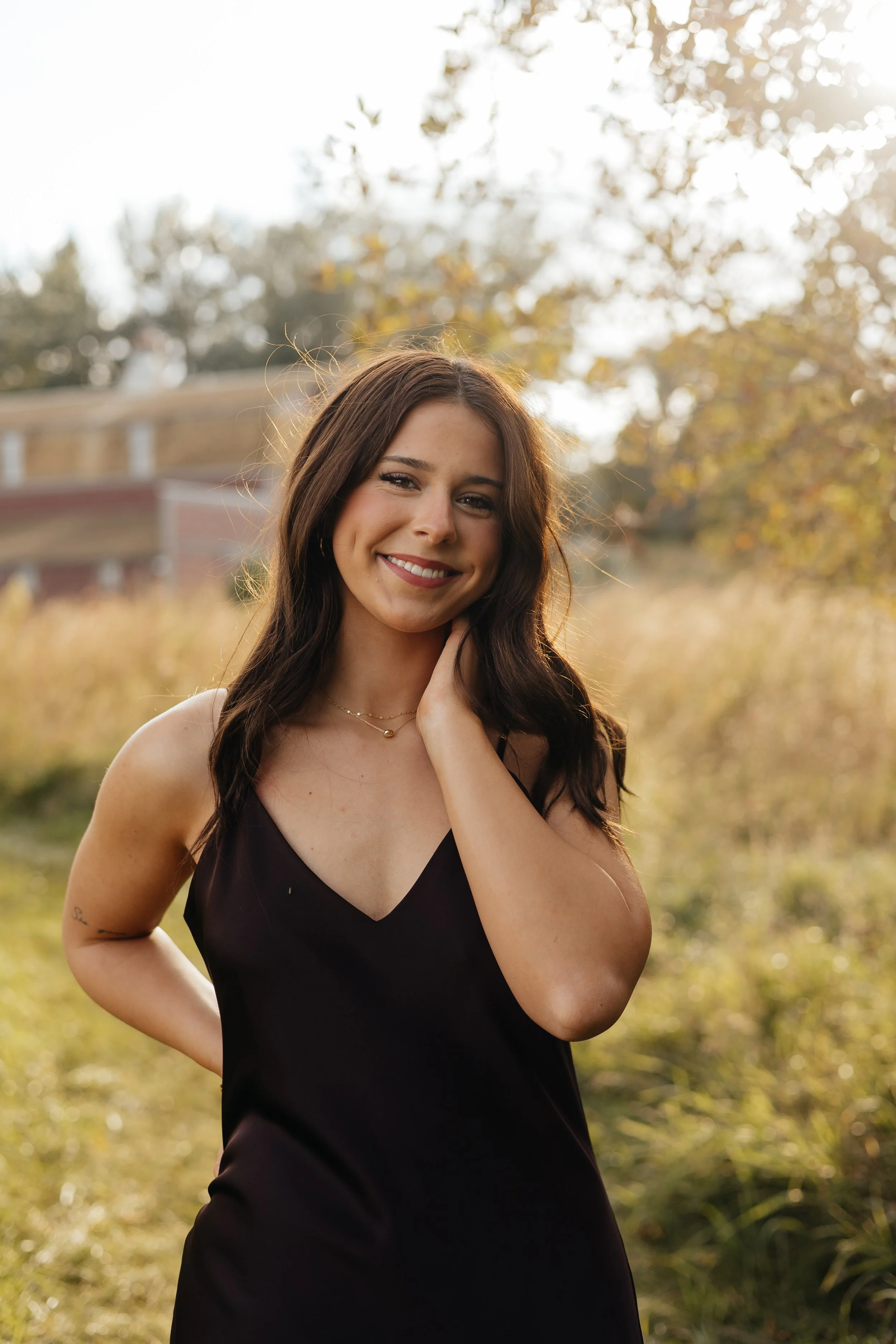 A young woman with long dark hair smiling outdoors in the golden sunlight, wearing a black dress and a delicate gold necklace, standing in a natural setting with trees and grass.