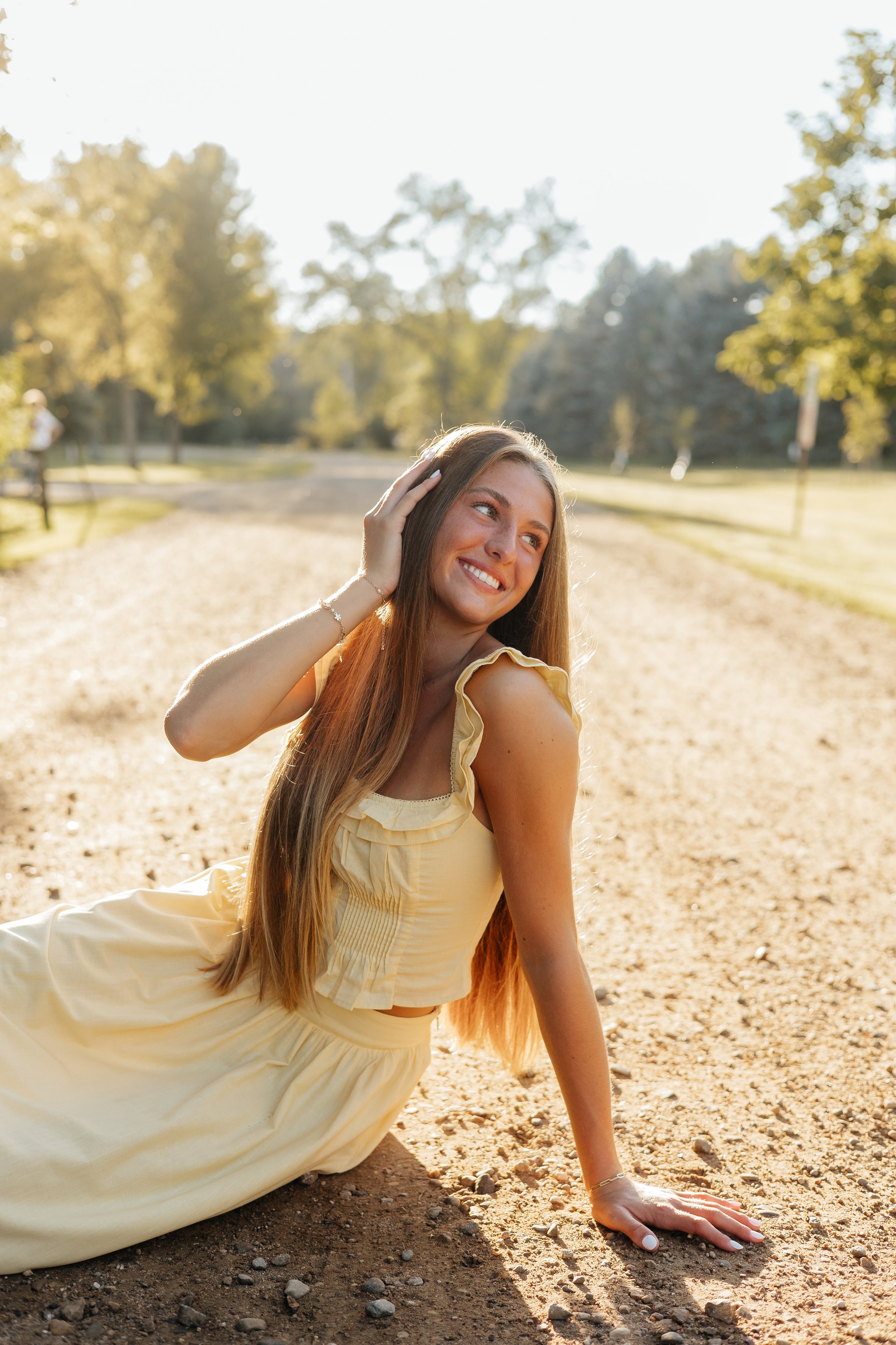 A young woman with long hair, wearing a yellow dress, is sitting on the ground in a park during sunlight, smiling and touching her hair.