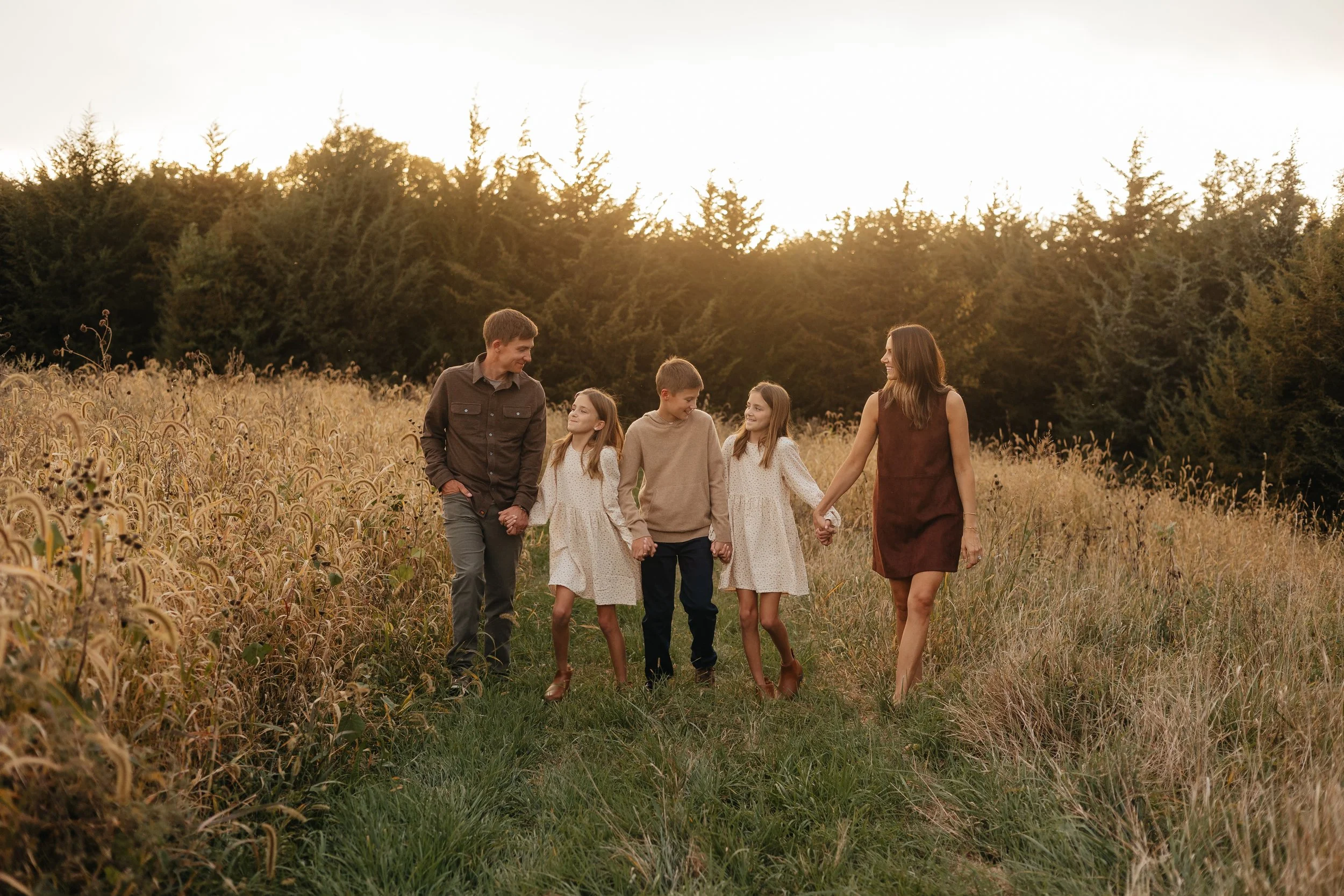 A family of six walking hand in hand through a grassy field at sunset, with trees in the background and smiling faces.
