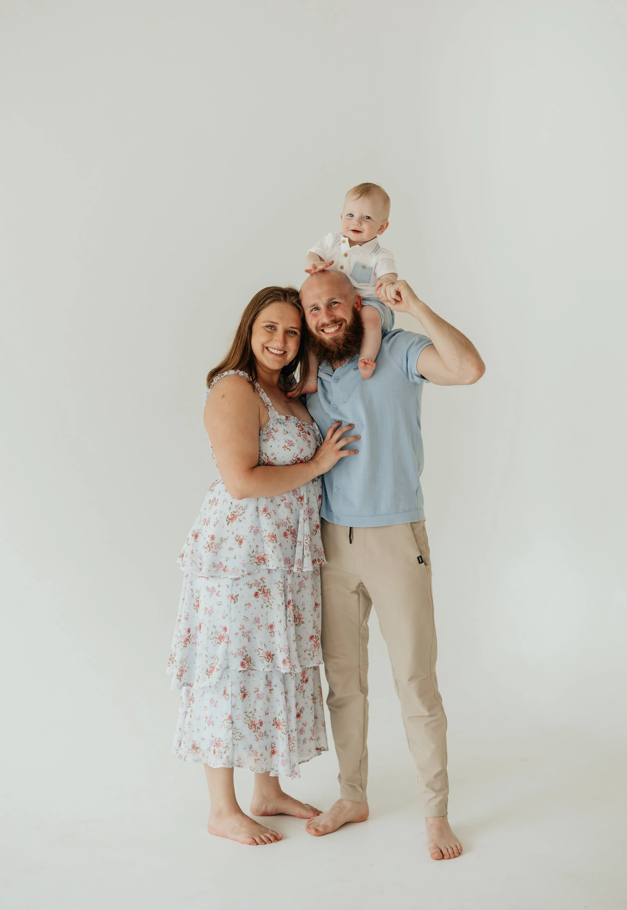 A happy family of three, a mother, father, and their baby, standing together against a plain white background. The baby is sitting on the father's shoulders, and everyone is smiling at the camera.