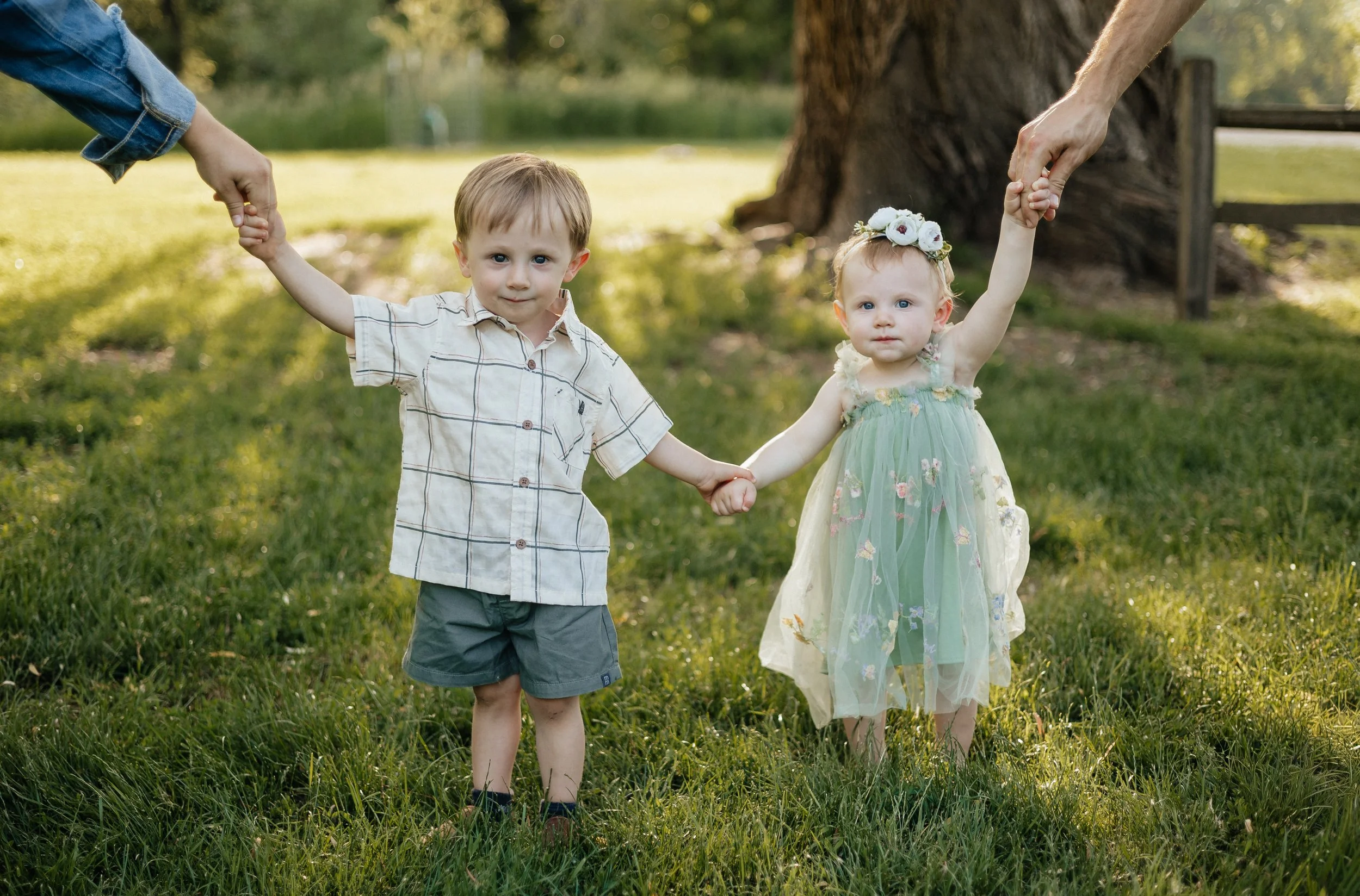 Two young children, a boy and a girl, holding hands with adults in a park, with green grass and trees in the background, during daylight.