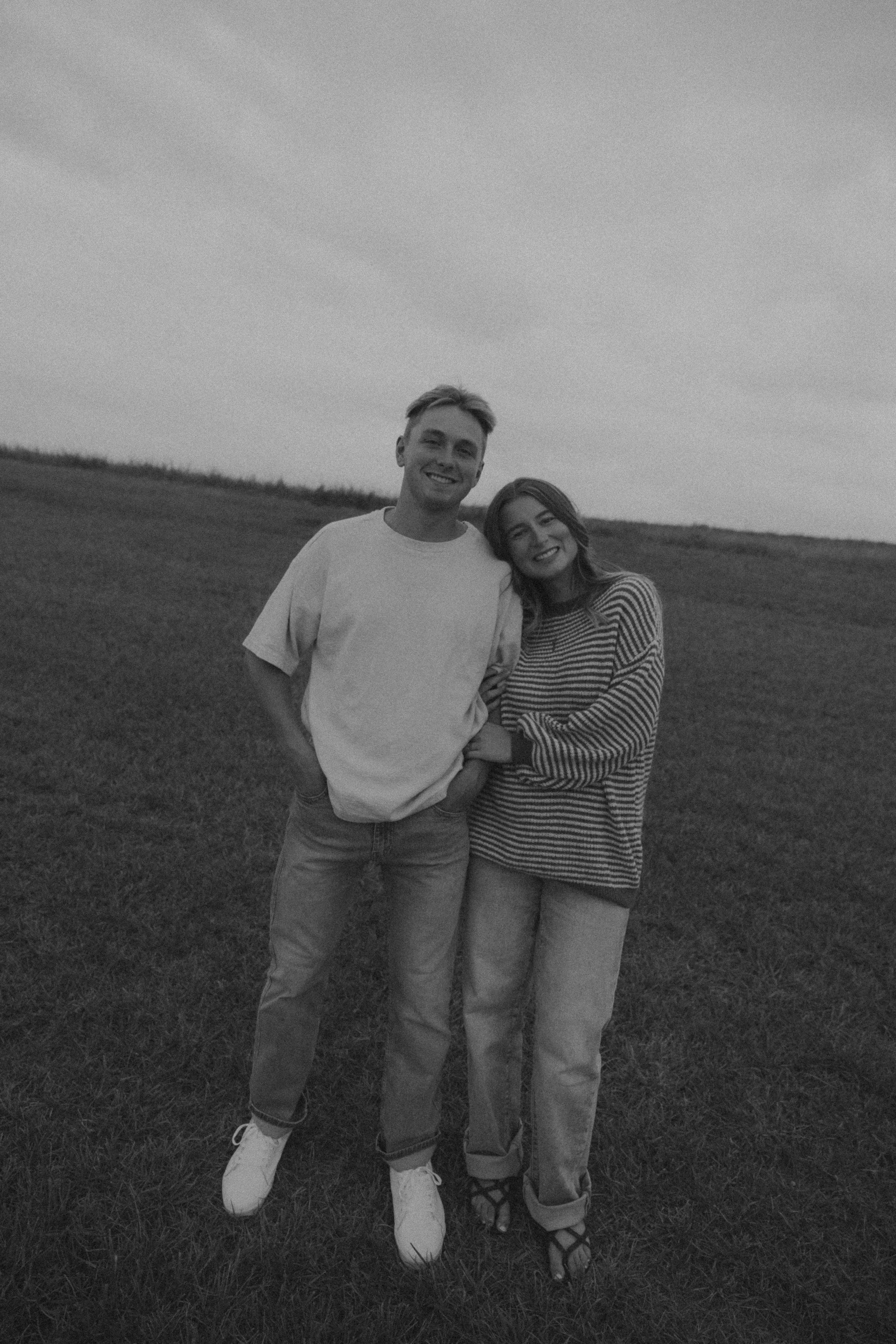 A black-and-white photo of a smiling young man and woman standing close together on a grassy field, with a cloudy sky in the background. The man is wearing a white t-shirt and jeans, and the woman is wearing a striped sweater and jeans.
