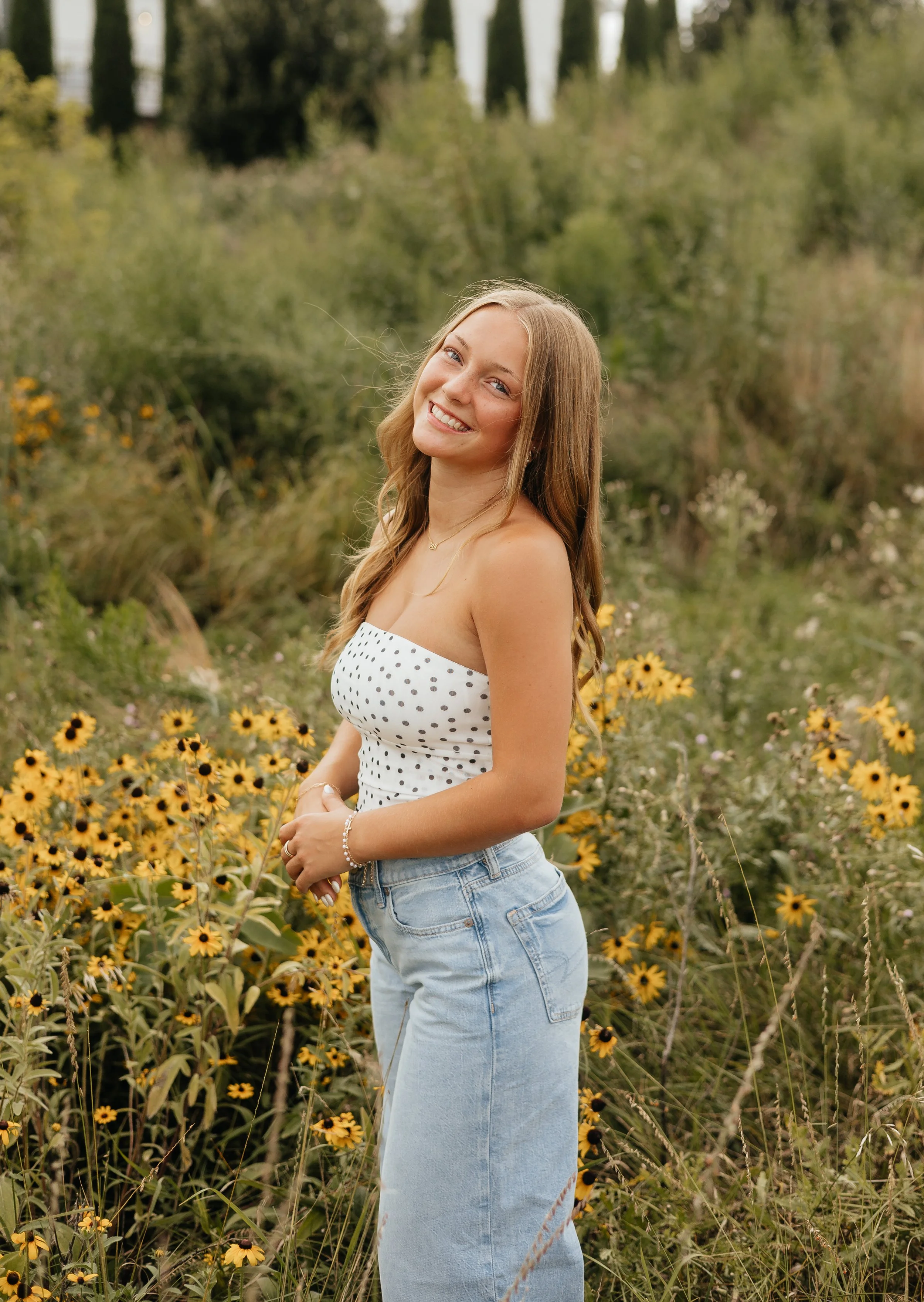 A young woman with long brown hair smiling and posing in a field of yellow flowers and greenery during daytime.