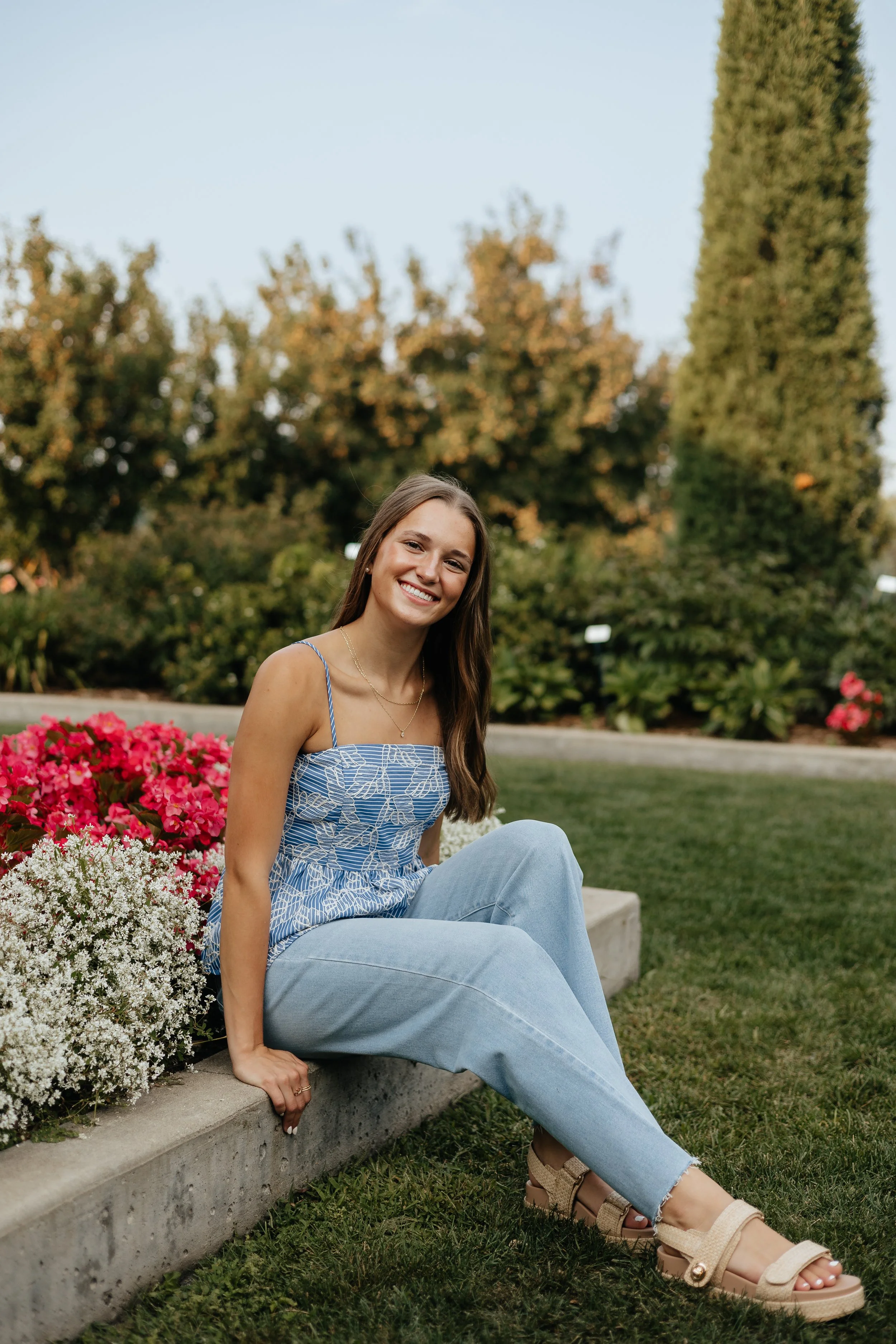 A young woman with long brown hair sitting on a concrete ledge in a park, smiling, wearing a blue patterned spaghetti strap top, light blue jeans, and beige sandals, with pink and white flowers in the foreground and tall green trees in the background