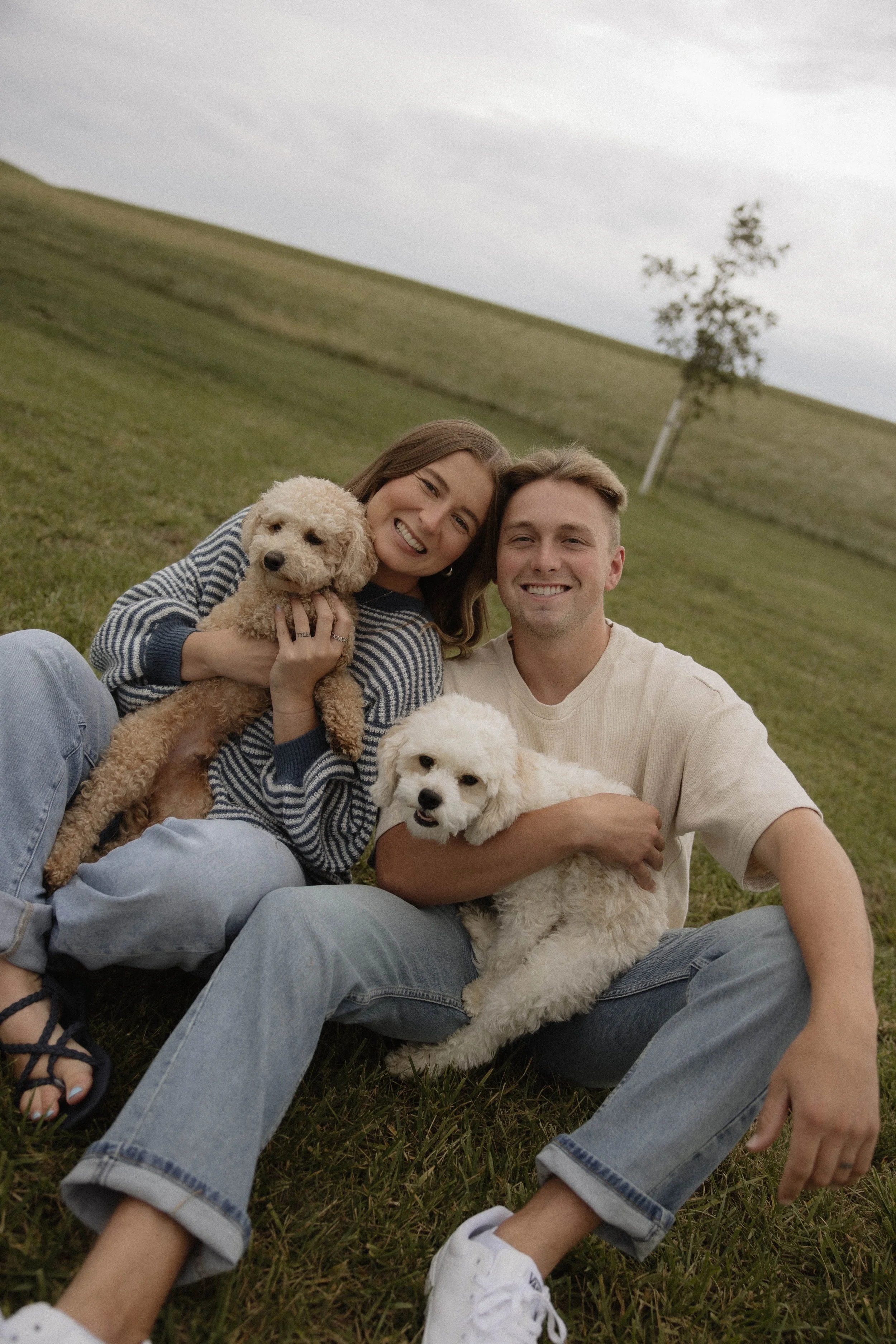 Young couple sitting on grass outdoors with two dogs, smiling at the camera, with a sloped grassy hill and a small tree in the background.