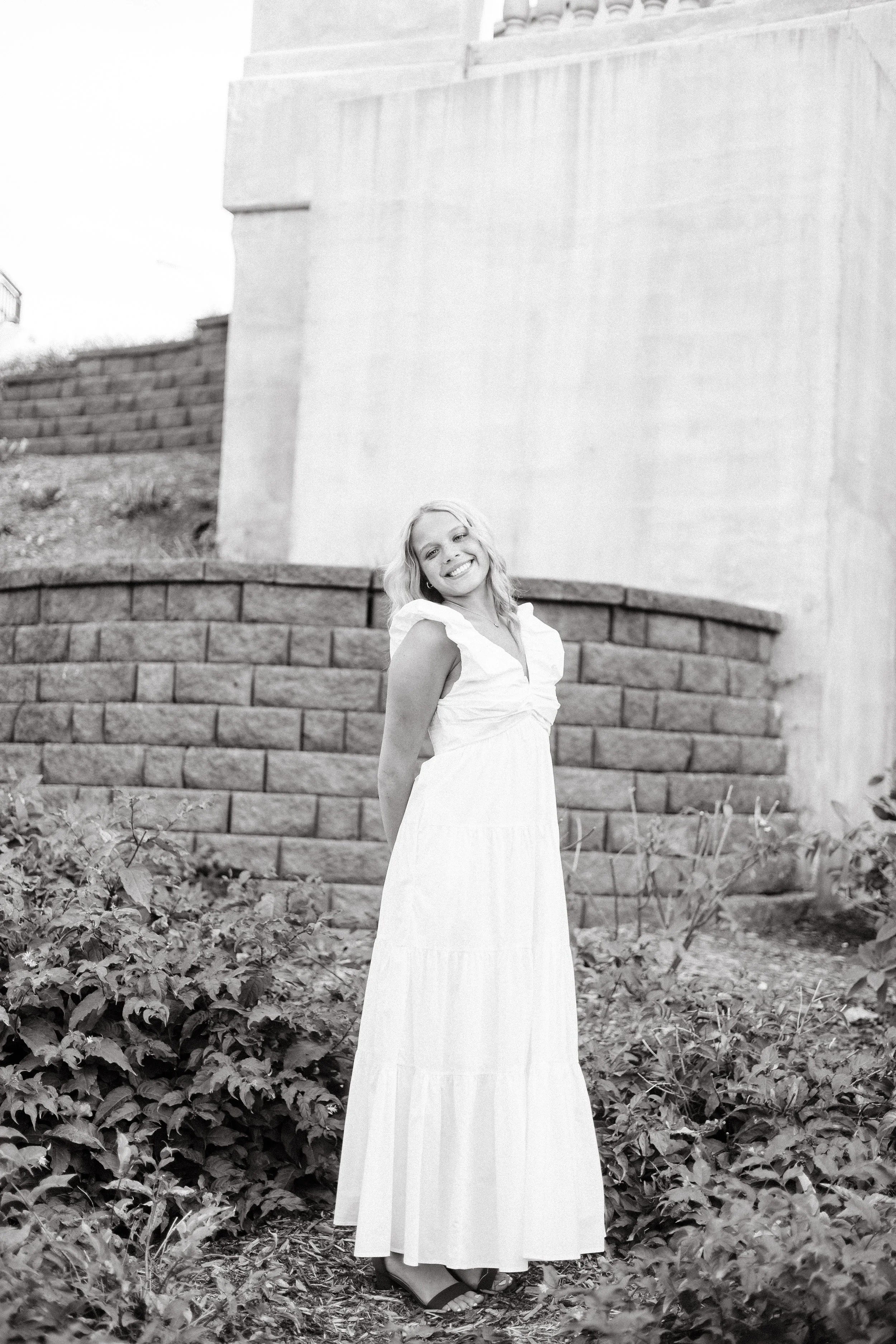 A woman with blonde hair wearing a sleeveless white dress and black sandals standing outdoors in front of a stone wall with plants at her feet, smiling at the camera.