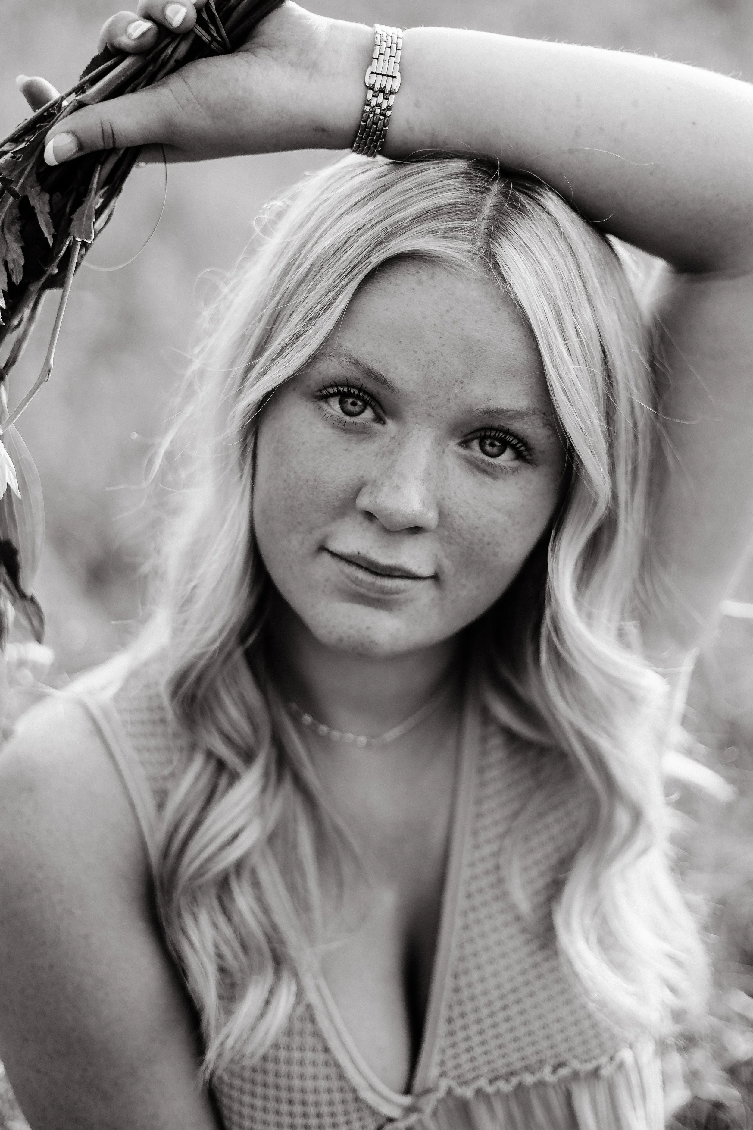 A close-up black and white photo of a young woman with long, wavy hair, looking at the camera with a slight smile. She is resting her arm on her head, wearing a watch and a sleeveless top.