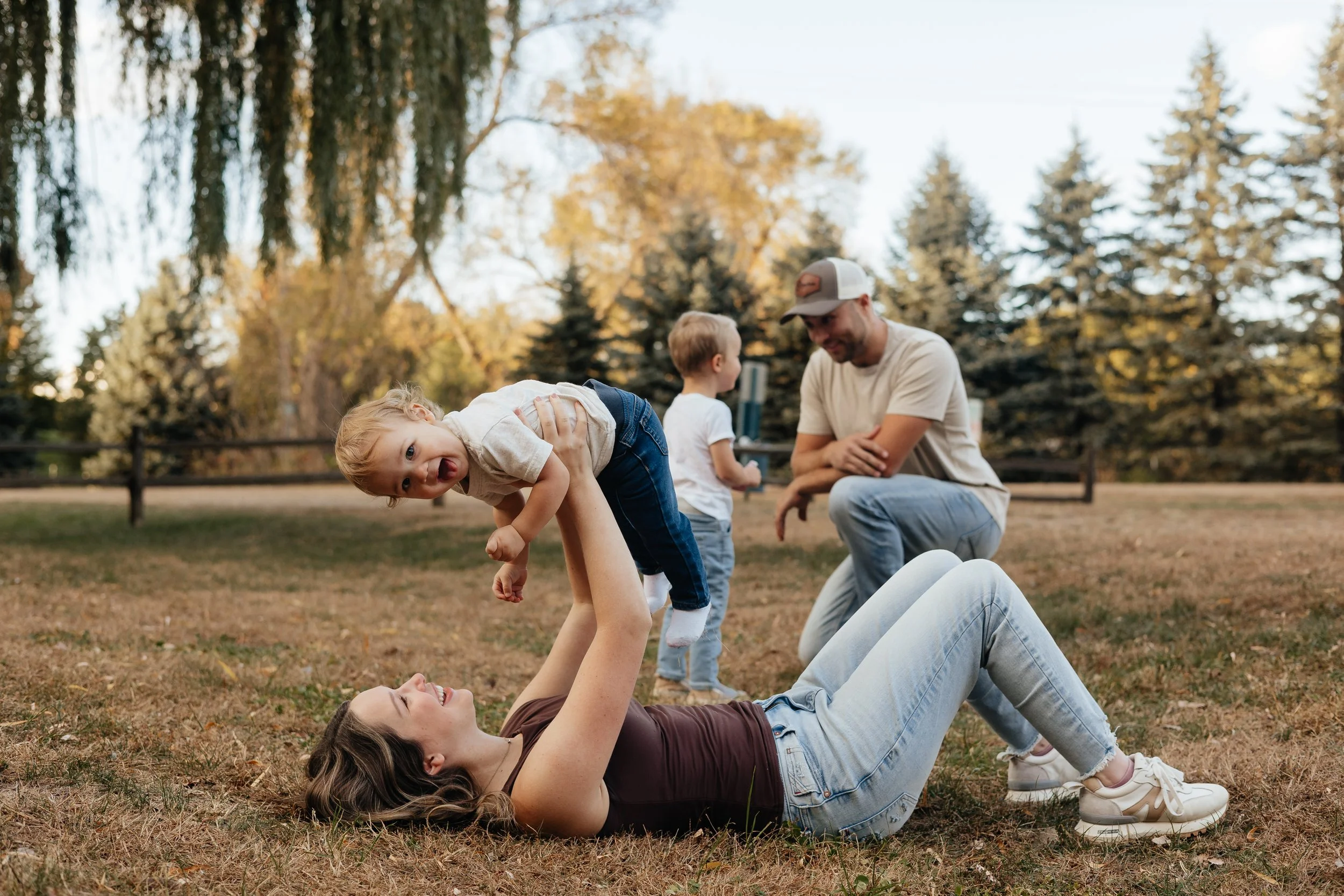 A woman lying on the grass playing with her children, a girl and a boy, in a park during the daytime. The girl is on the woman's legs, and the woman is holding her hands while smiling. The boy is standing nearby, and another girl is being held in the