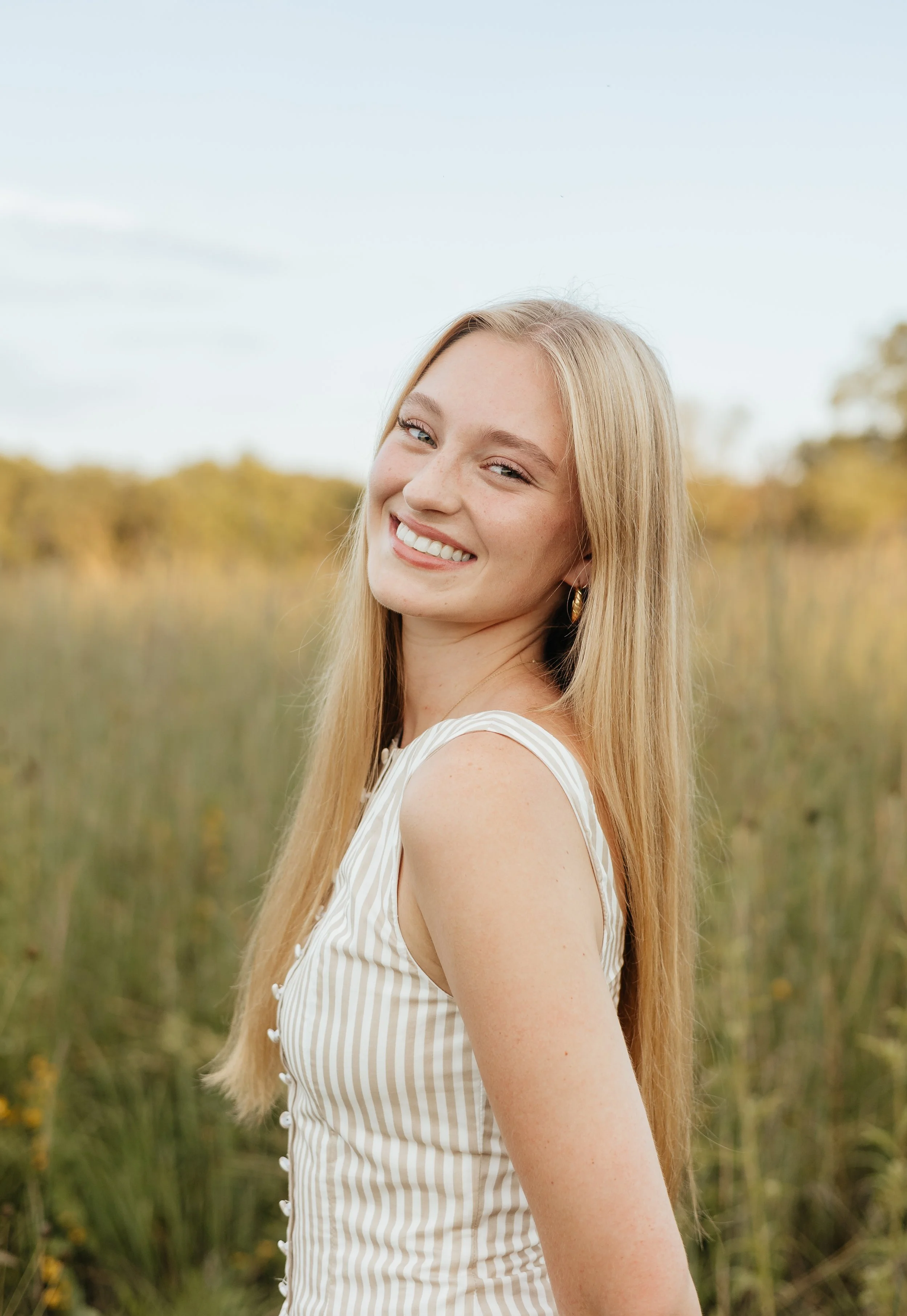 A young woman with long blonde hair smiling outdoors in a grassy field with trees in the background.