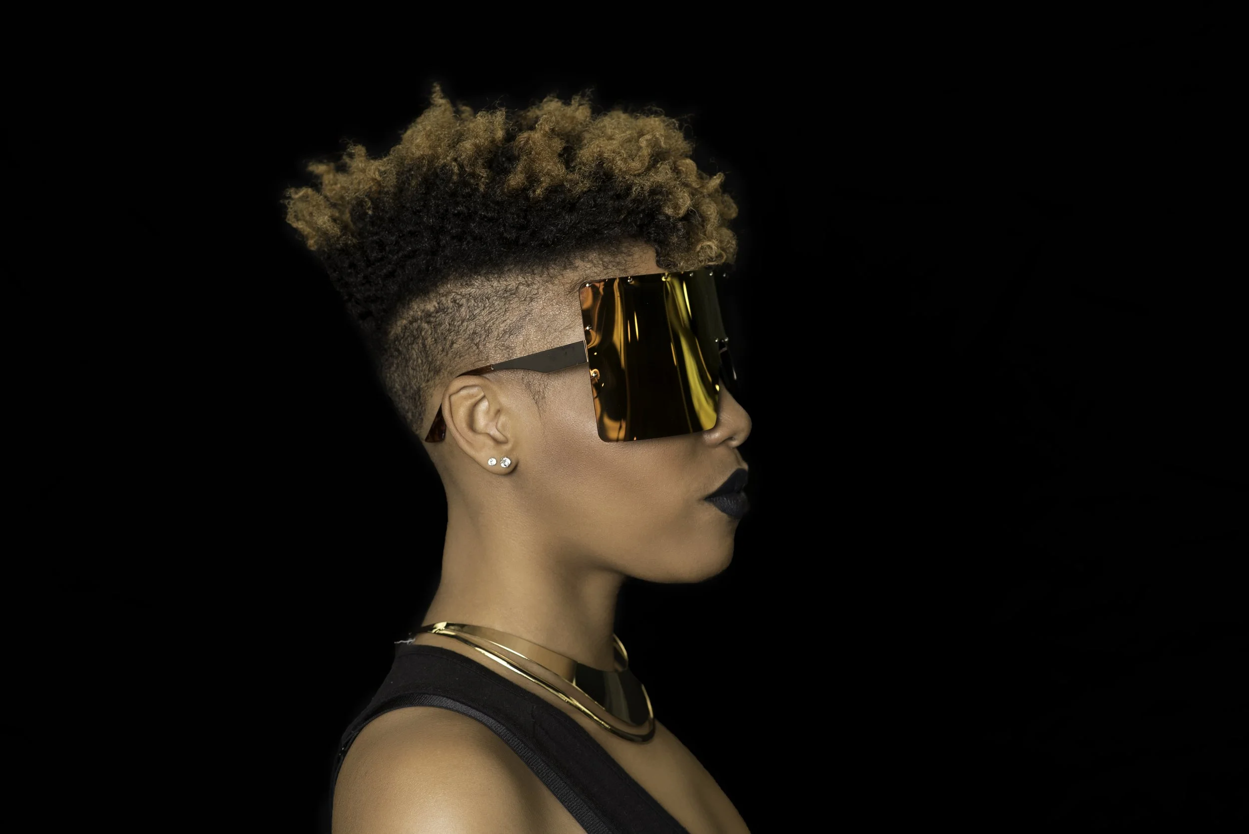 Side profile of a woman with short, curly hair with a fade on the sides, wearing large, reflective sunglasses, gold jewelry, and black lipstick against a black background.