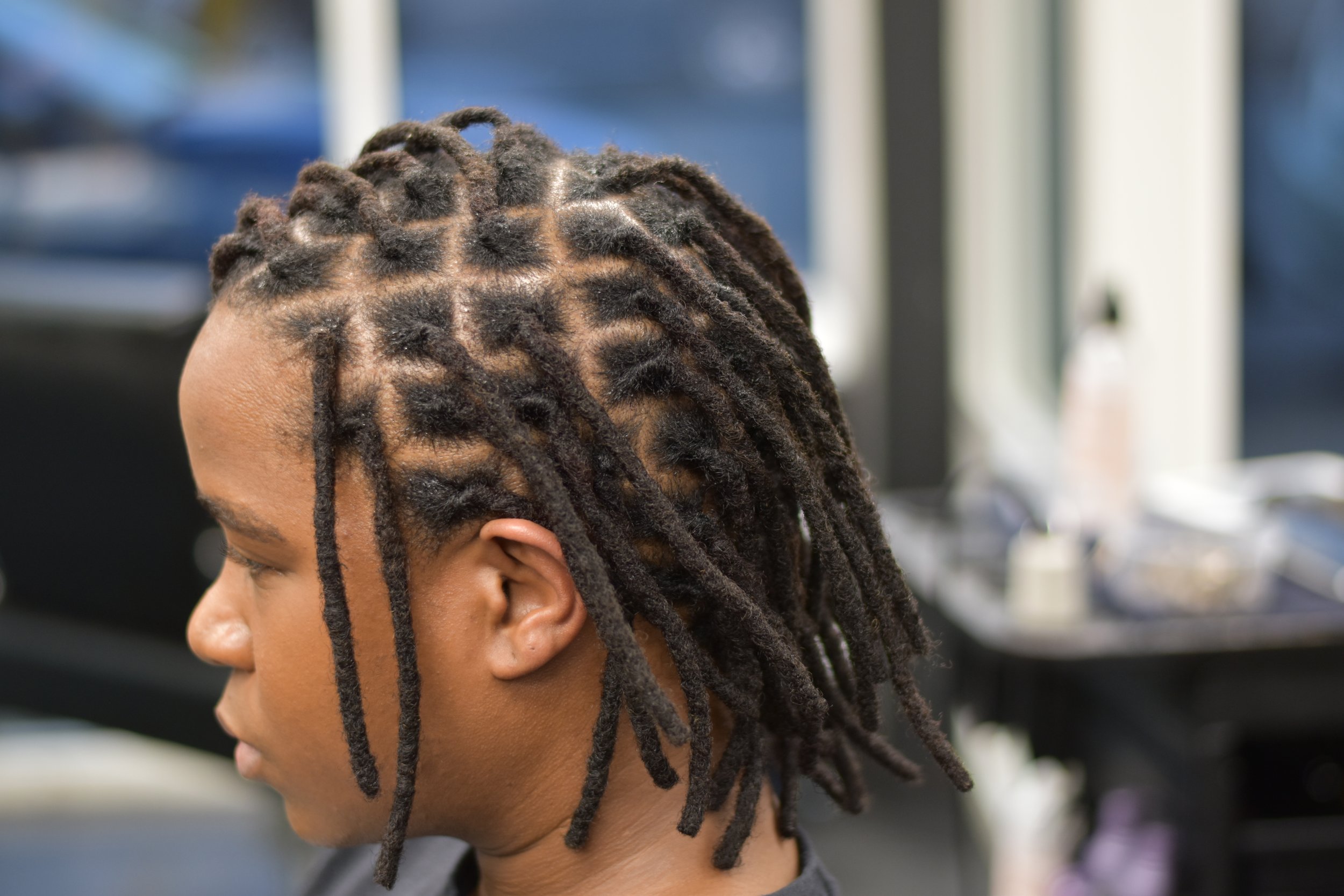 Side profile of a man with neat, professional locs sitting in a salon chair, featuring a blurred background of the Mr. Naturalz Salon interior.
