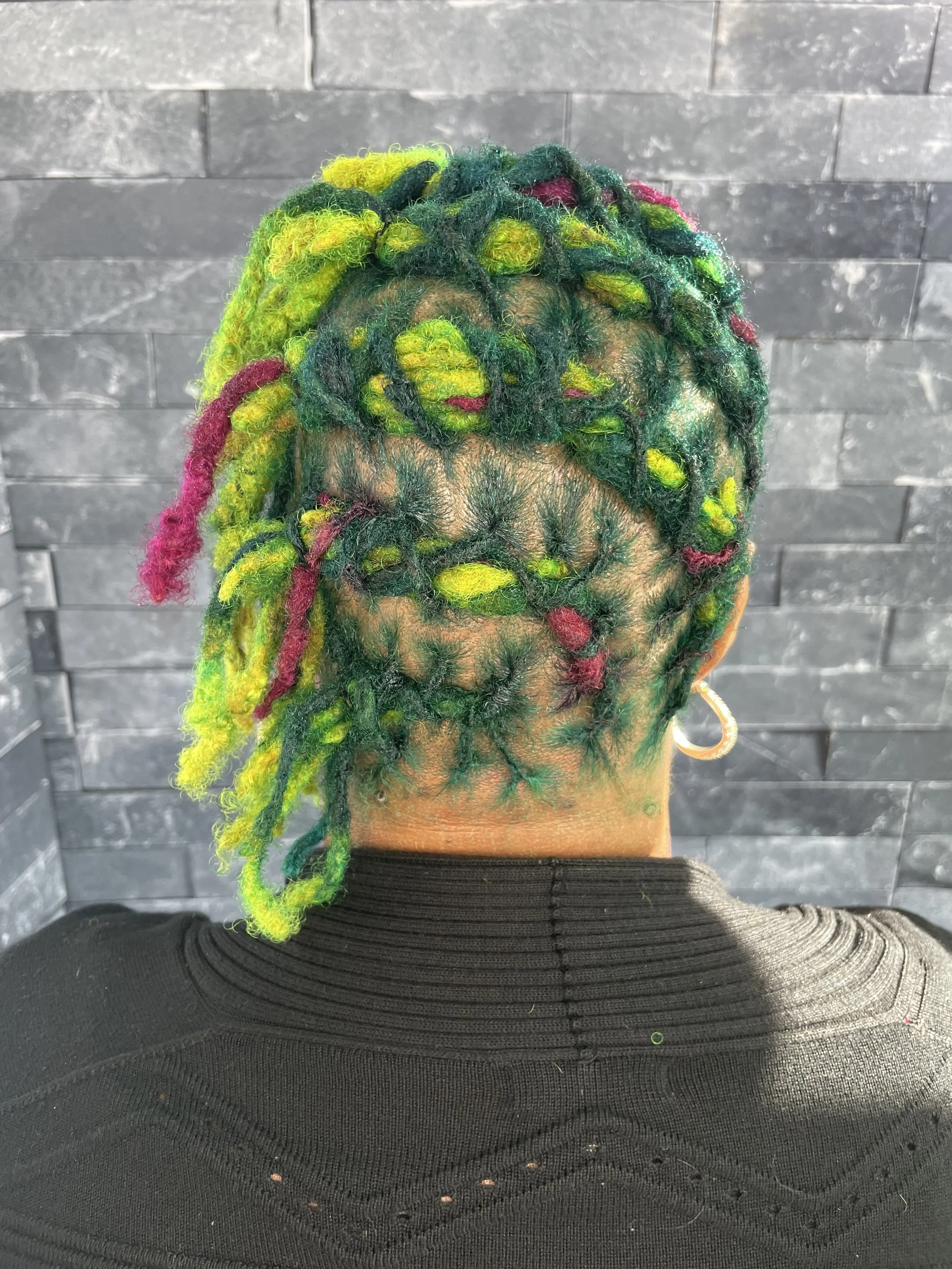 Back of a person's head with multicolored dreadlocks in green, yellow, pink, and dark shades, set against a dark brick wall background.