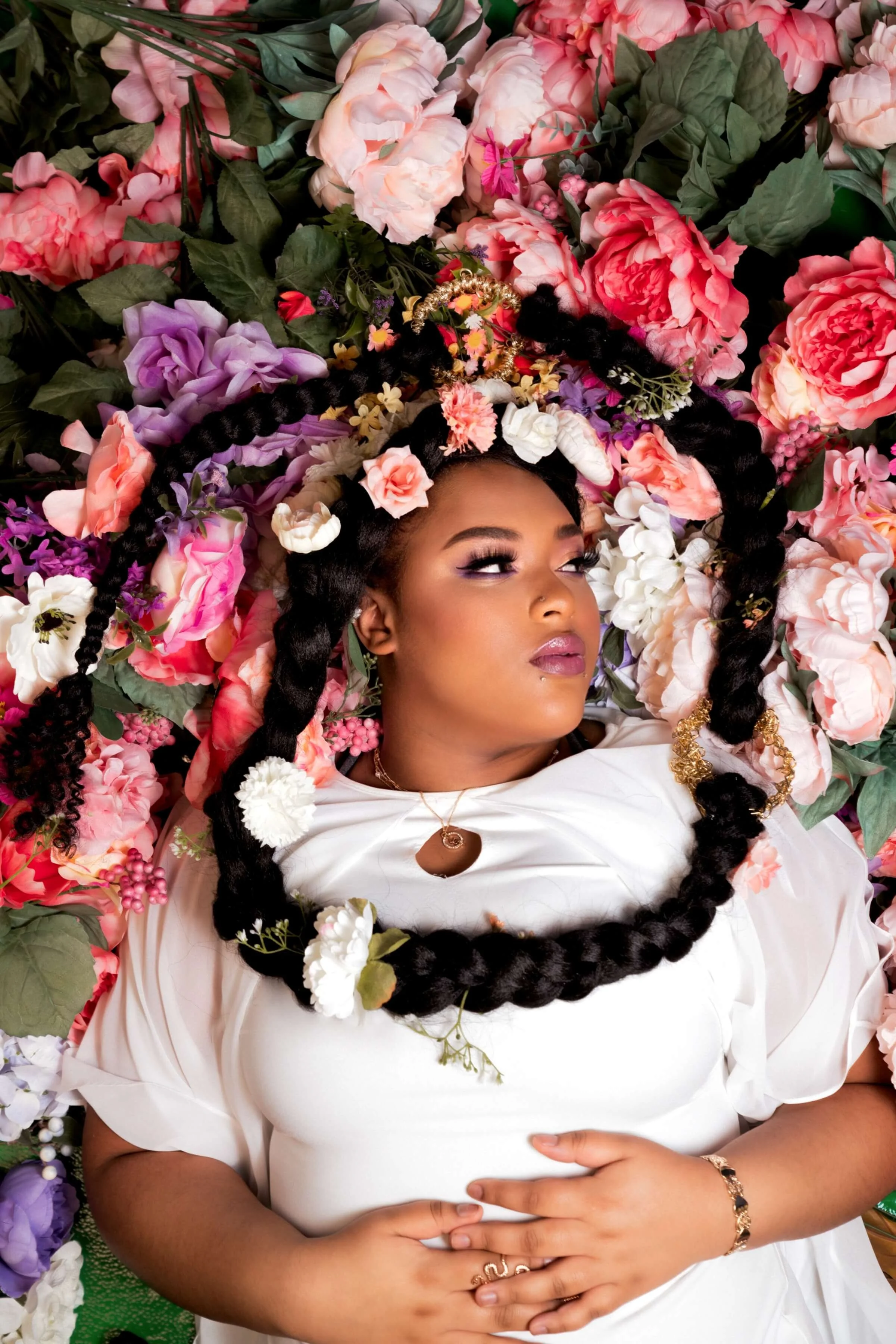 Artistic overhead view of a woman in a white dress lying among colorful flowers with long braids decorated with floral accents and gold jewelry.