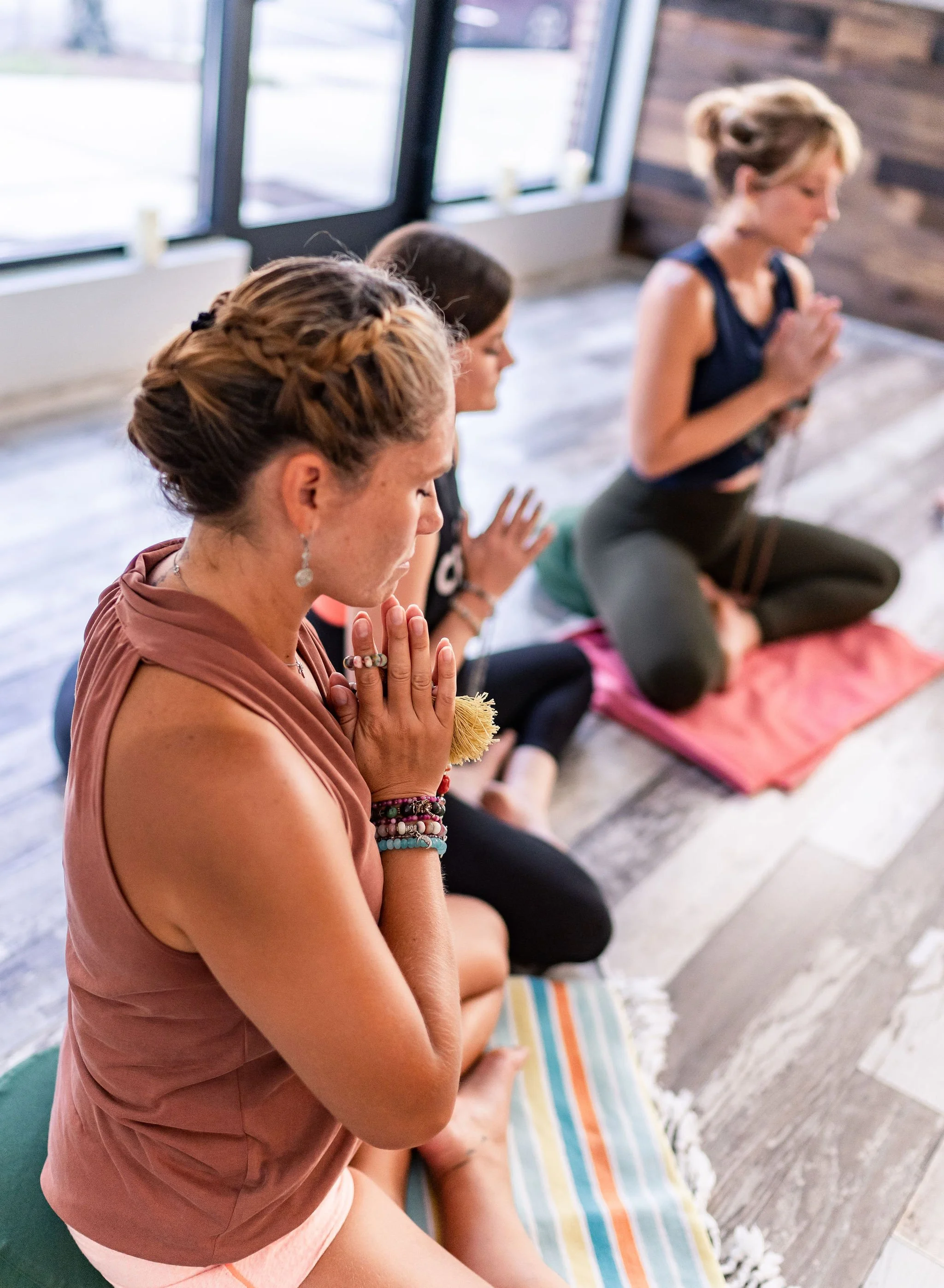Women practicing yoga or meditation, seated on mats indoors near large windows, with hands in prayer position and eyes closed.