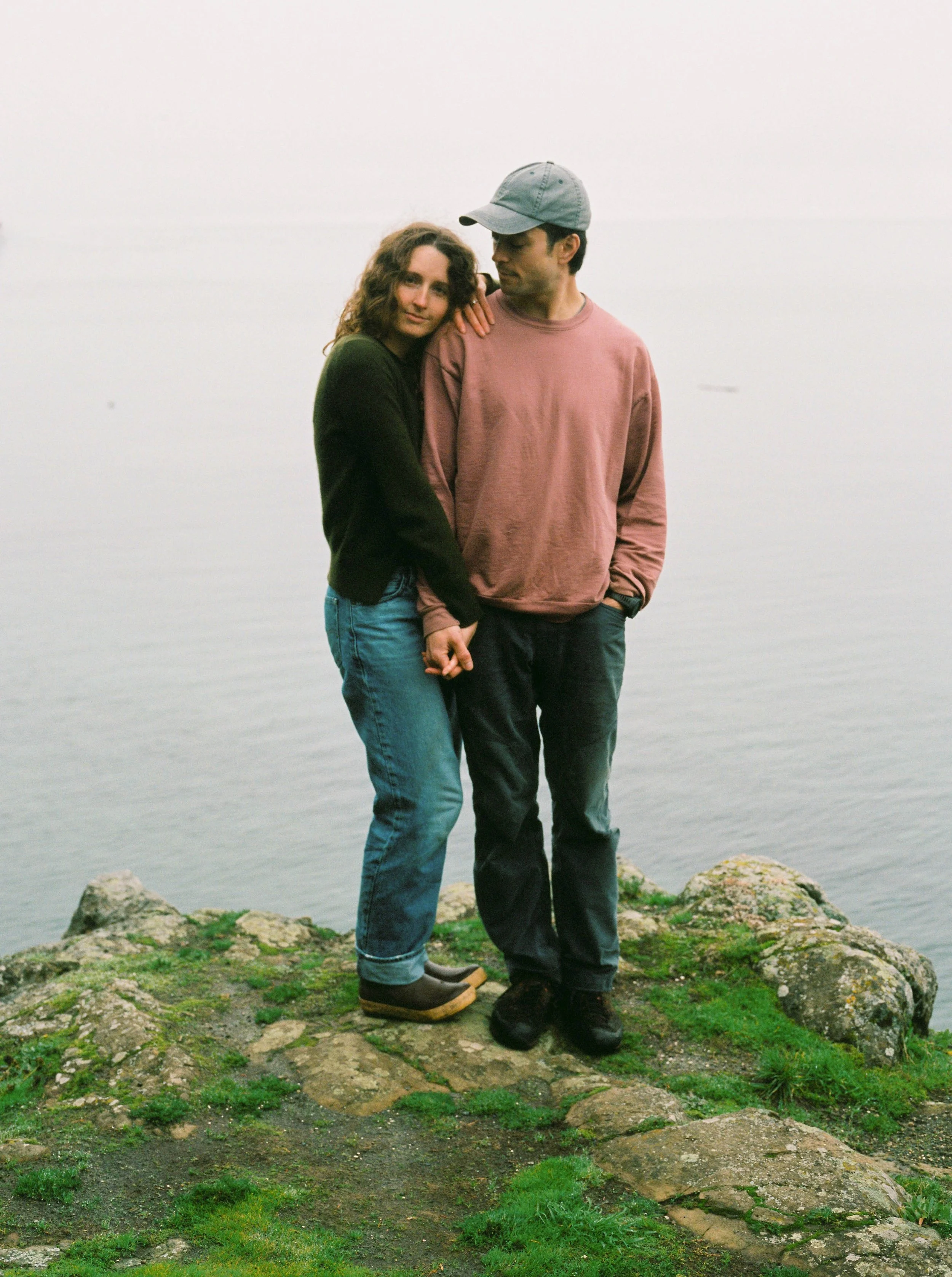 A man and woman walking on rocks near a body of water. The woman is leaning her head on the man's shoulder and holding his hand, both dressed casually in jeans and sweatshirts, with the woman wearing boots and the man wearing a cap.