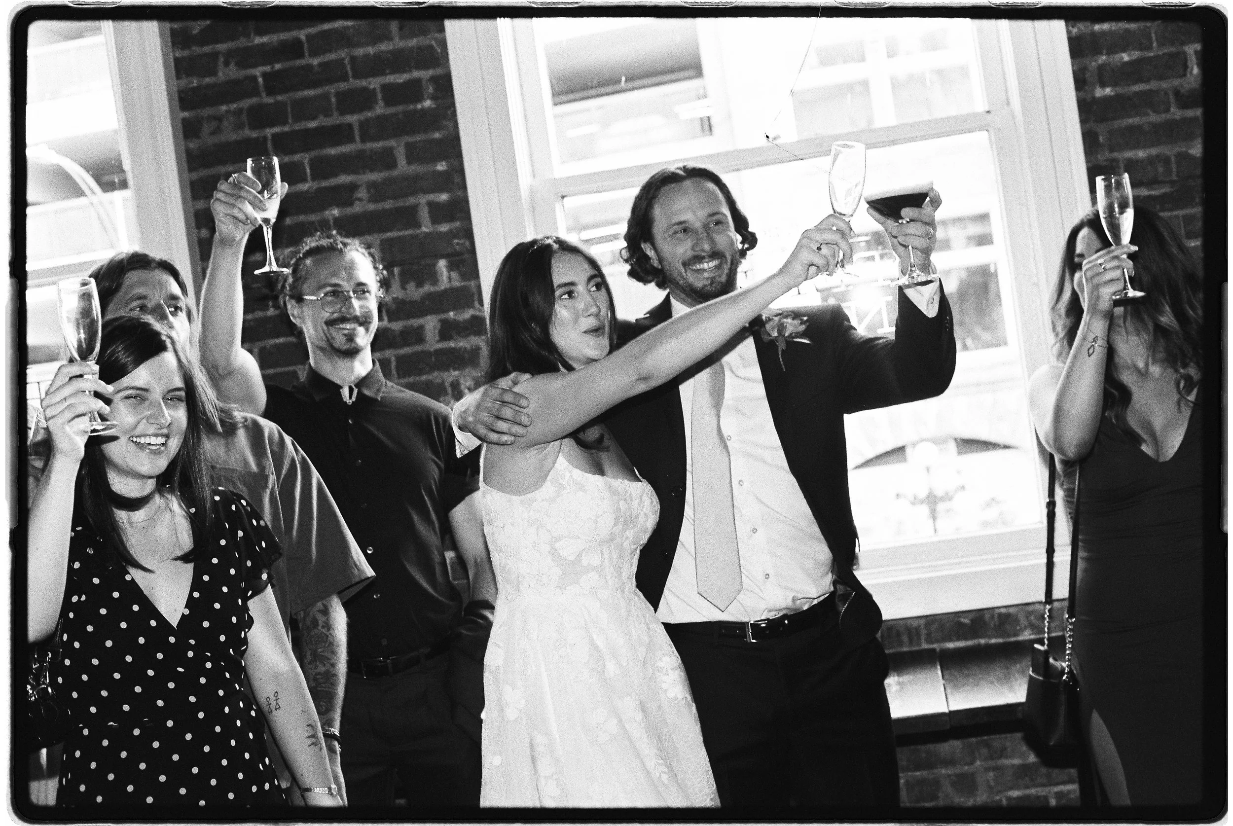 A group of people celebrating with champagne toasts at a wedding reception. The bride and groom are in the center, smiling and holding glasses, with friends around them.