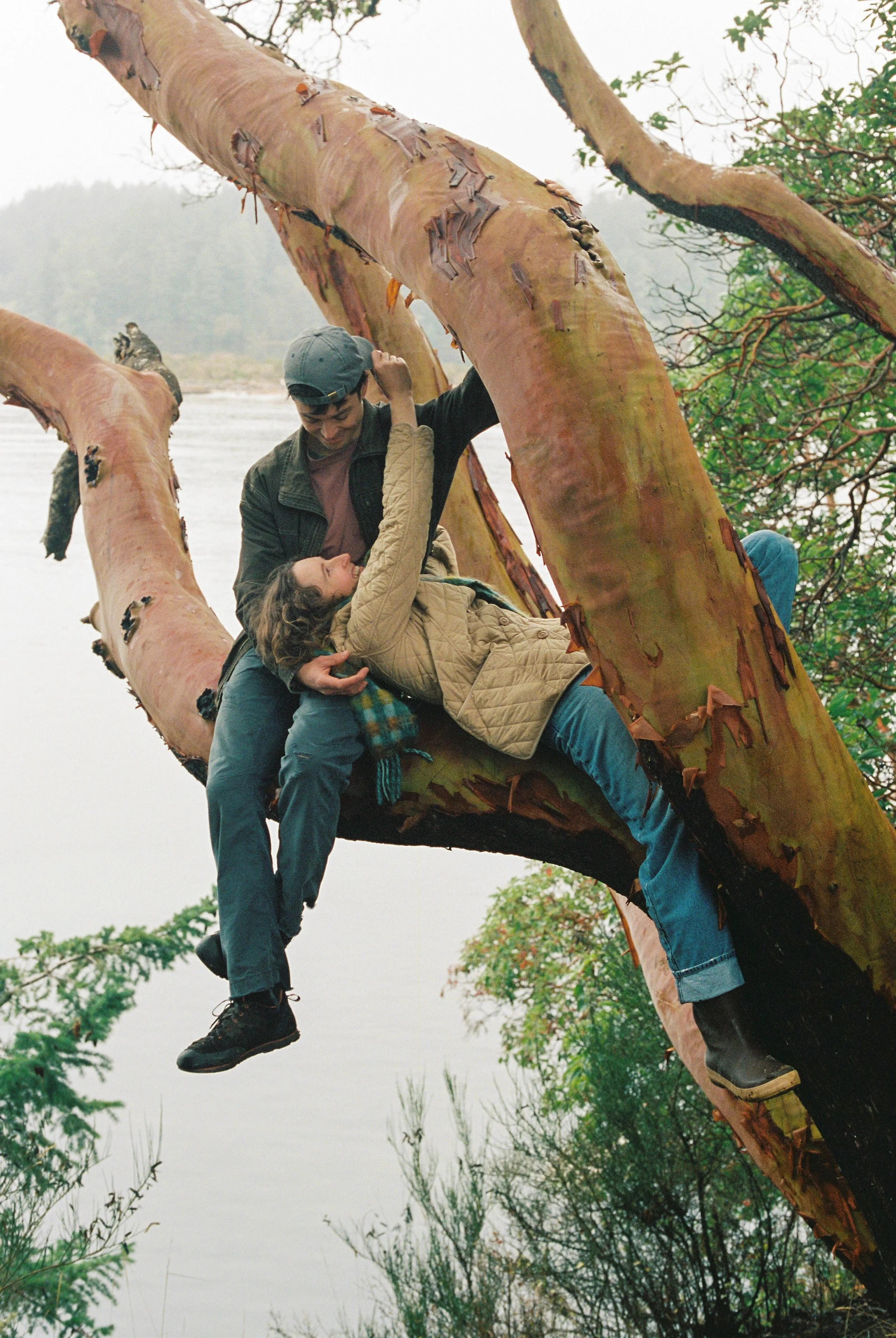 A young couple sits on a bend of a large tree branch over a river, the woman leaning back and smiling as the man supports her. They are outdoors surrounded by trees and cloudy sky.