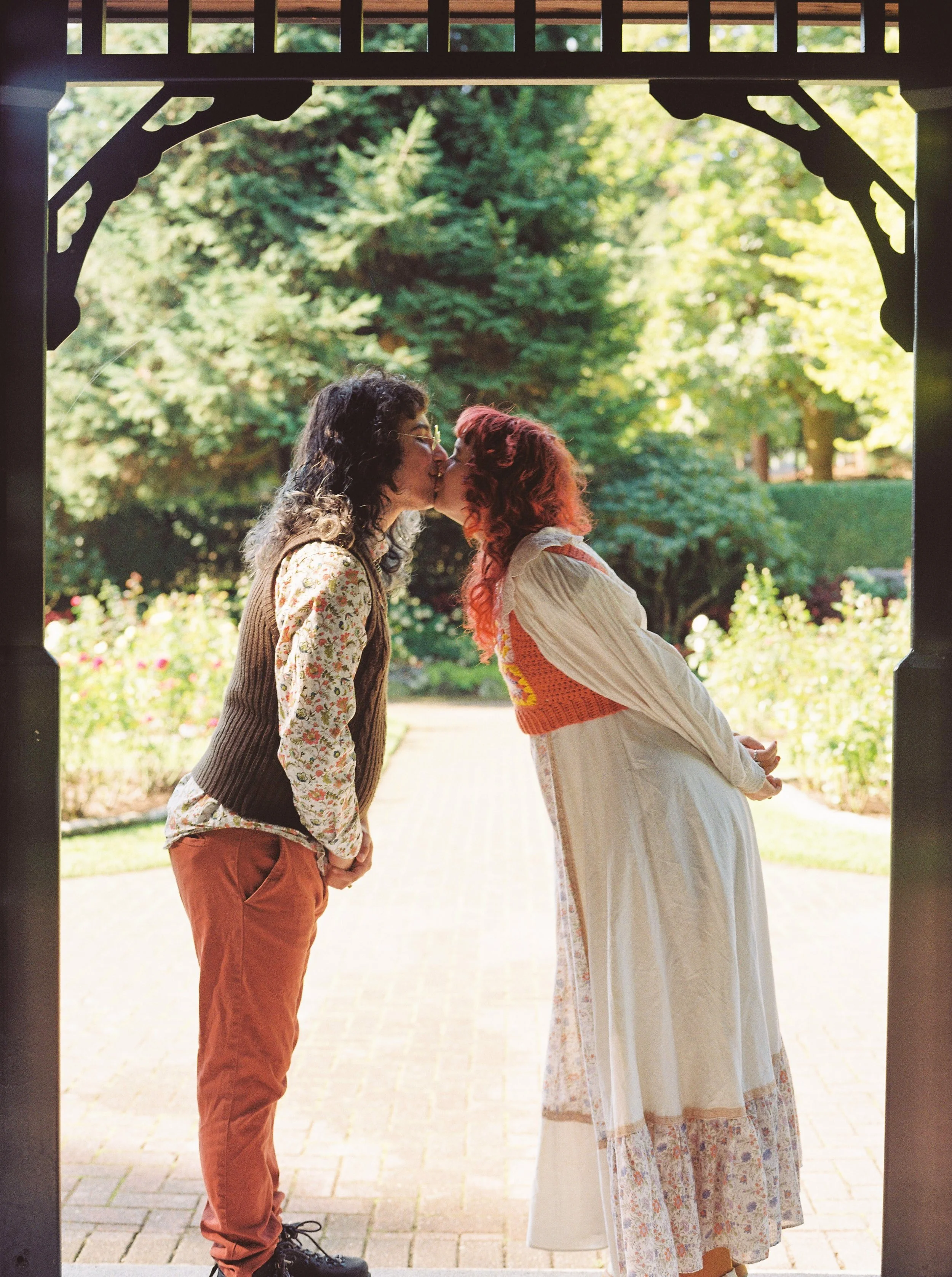 Two women stand close under a gazebo, sharing a kiss in a garden with trees and flowers in the background.