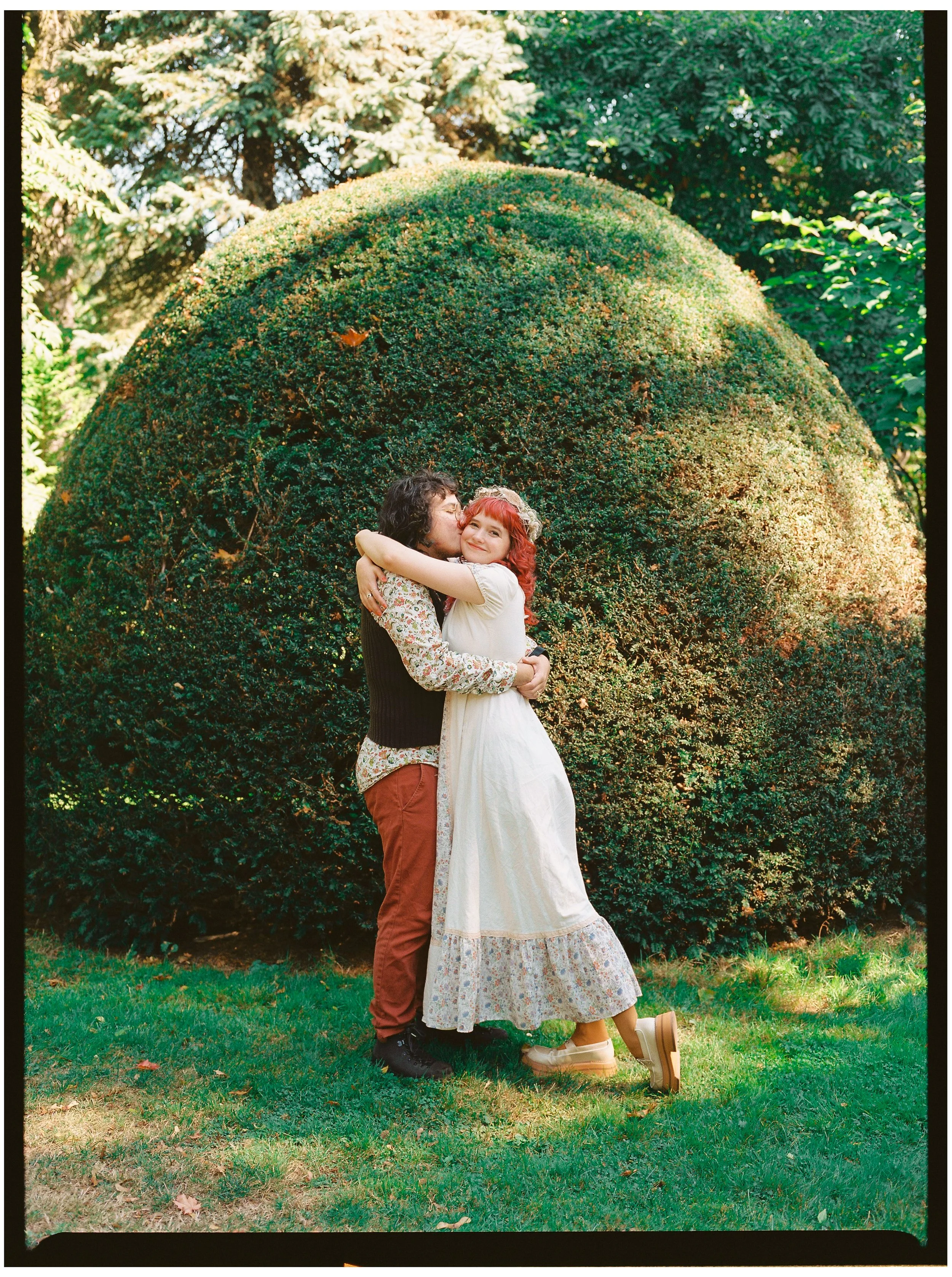 A man and woman embracing in front of a large, rounded shrub inside a lush garden, with tall trees in the background, during daytime.