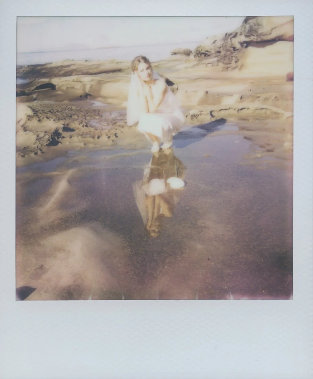 A woman sitting on a rock at the beach with her reflection visible in the wet sand, surrounded by rocks and water, during sunset or sunrise.