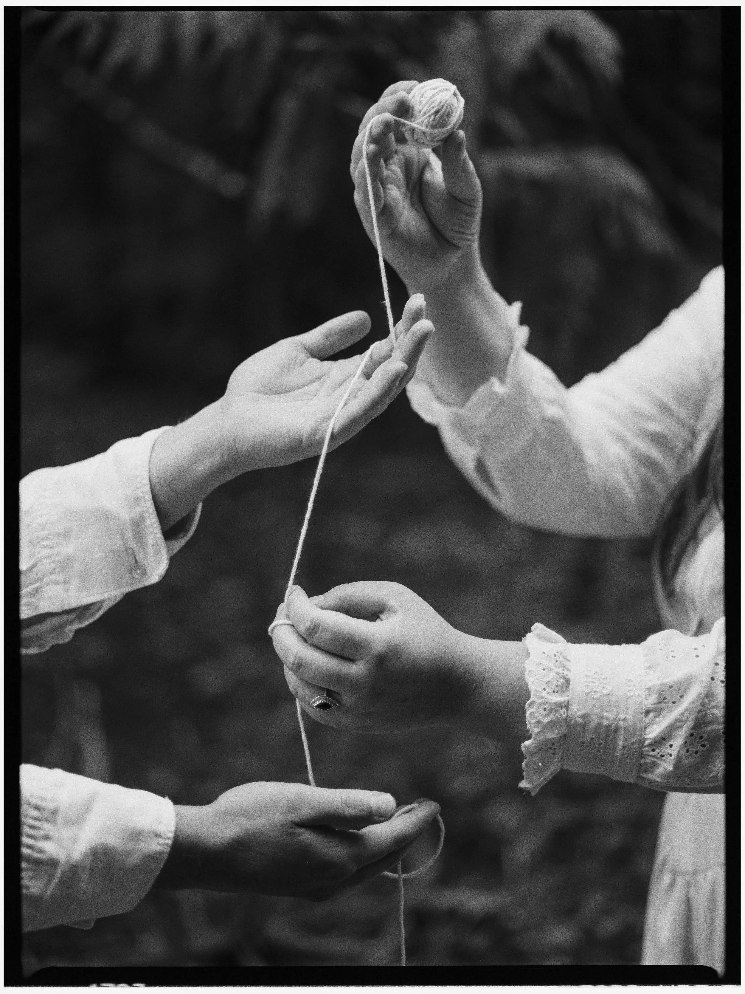 Three pairs of hands are involved in knitting or crocheting with yarn. The hands are outdoors, and the background is blurred.