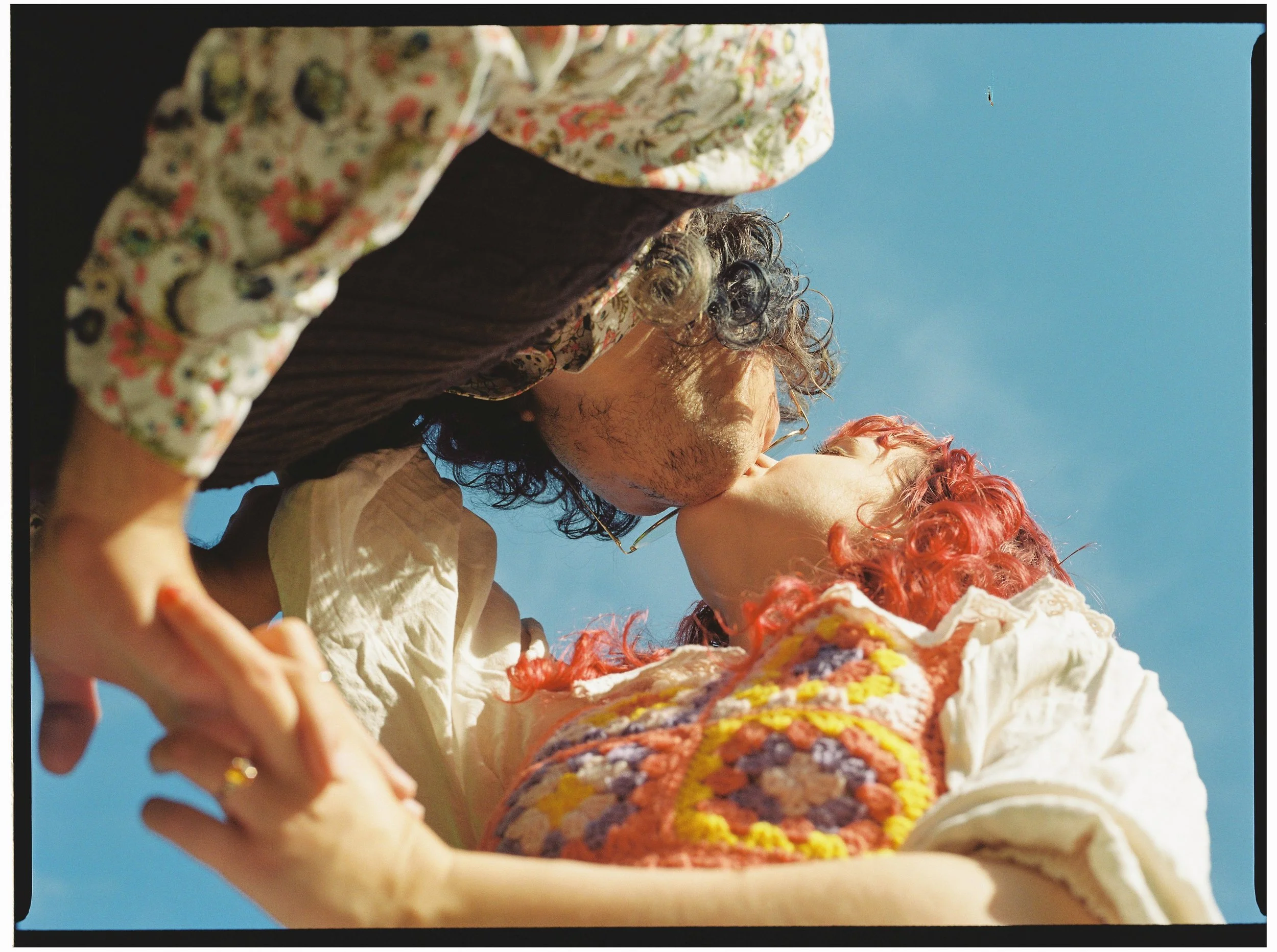 A couple kissing outdoors against a blue sky, both with curly hair, the woman has red hair and is wearing a colorful knitted top, the man has dark hair and is wearing a floral shirt and brown vest, holding hands.