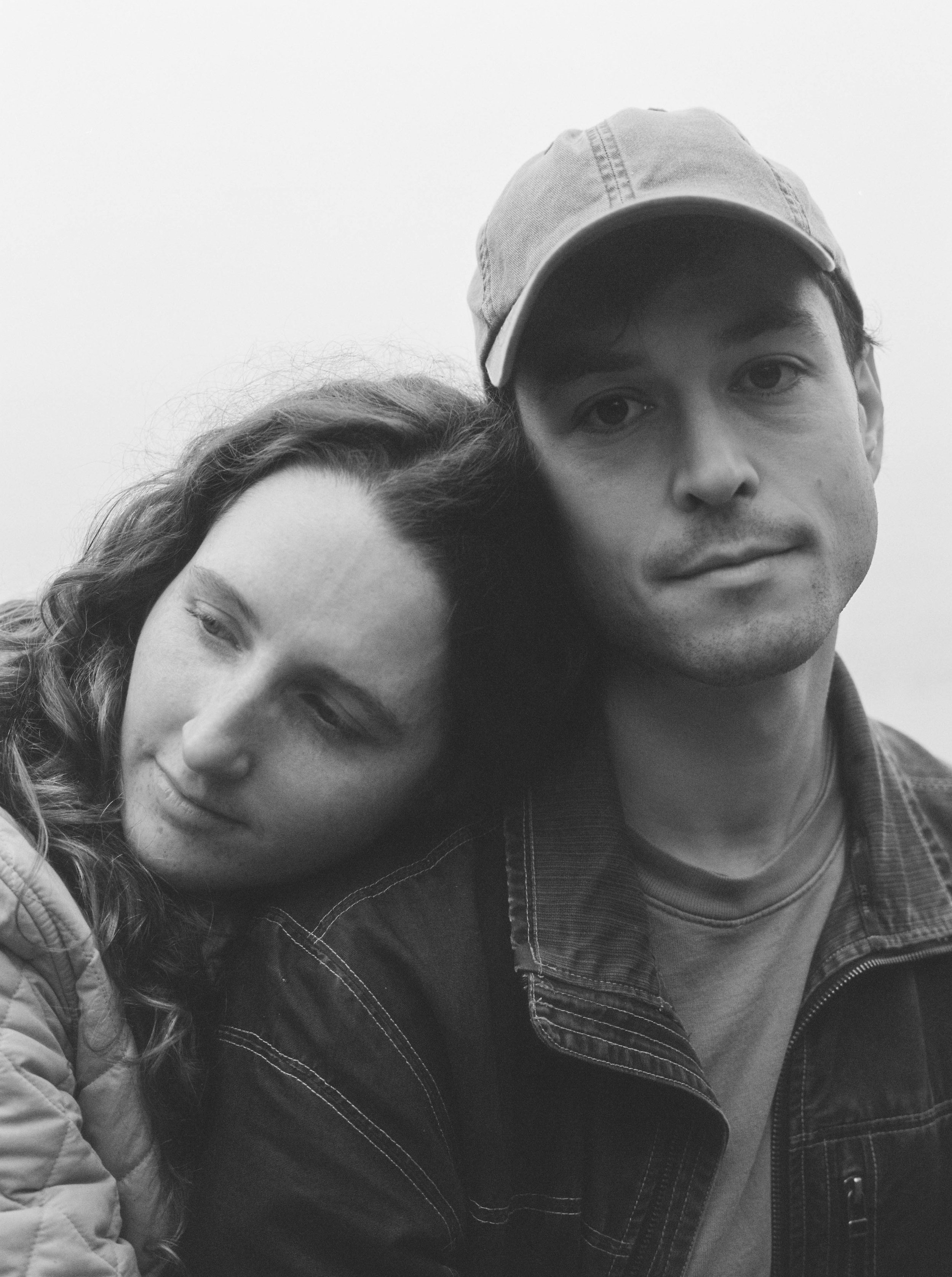A black-and-white photo of a man and woman close together, with the woman resting her head on the man's shoulder. The man is wearing a cap and a denim jacket, and the woman is wearing a quilted jacket. Both have neutral expressions.
