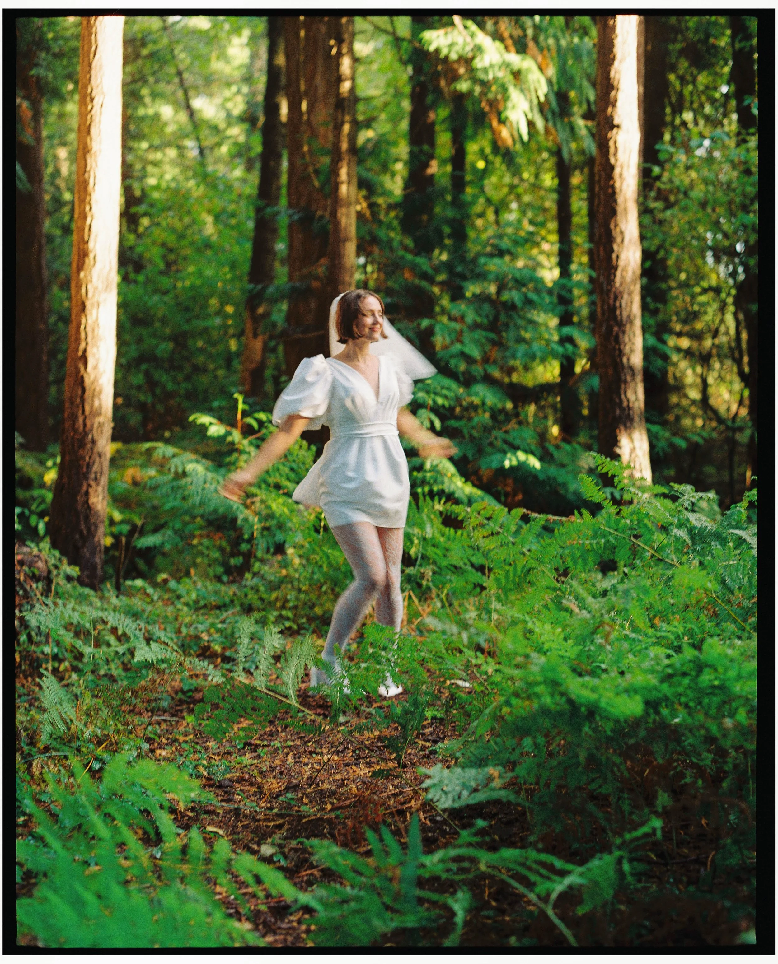 A woman in a white dress with puffed sleeves and lace tights is joyfully walking through a forest with tall trees and green foliage.
