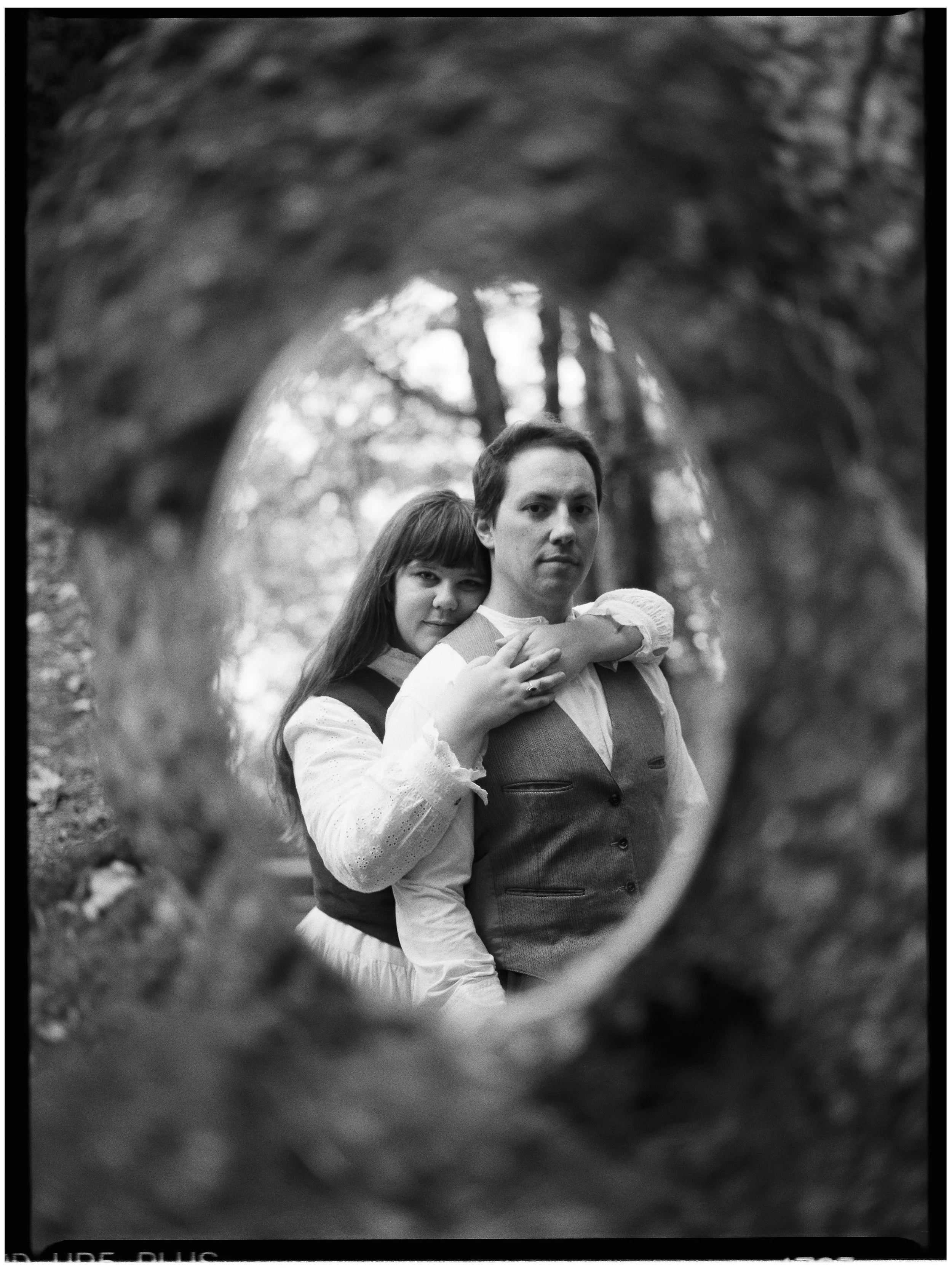 A black and white photo of a couple, seen through a round opening in a tree trunk, with the woman resting her head on the man's shoulder and both looking at the camera.
