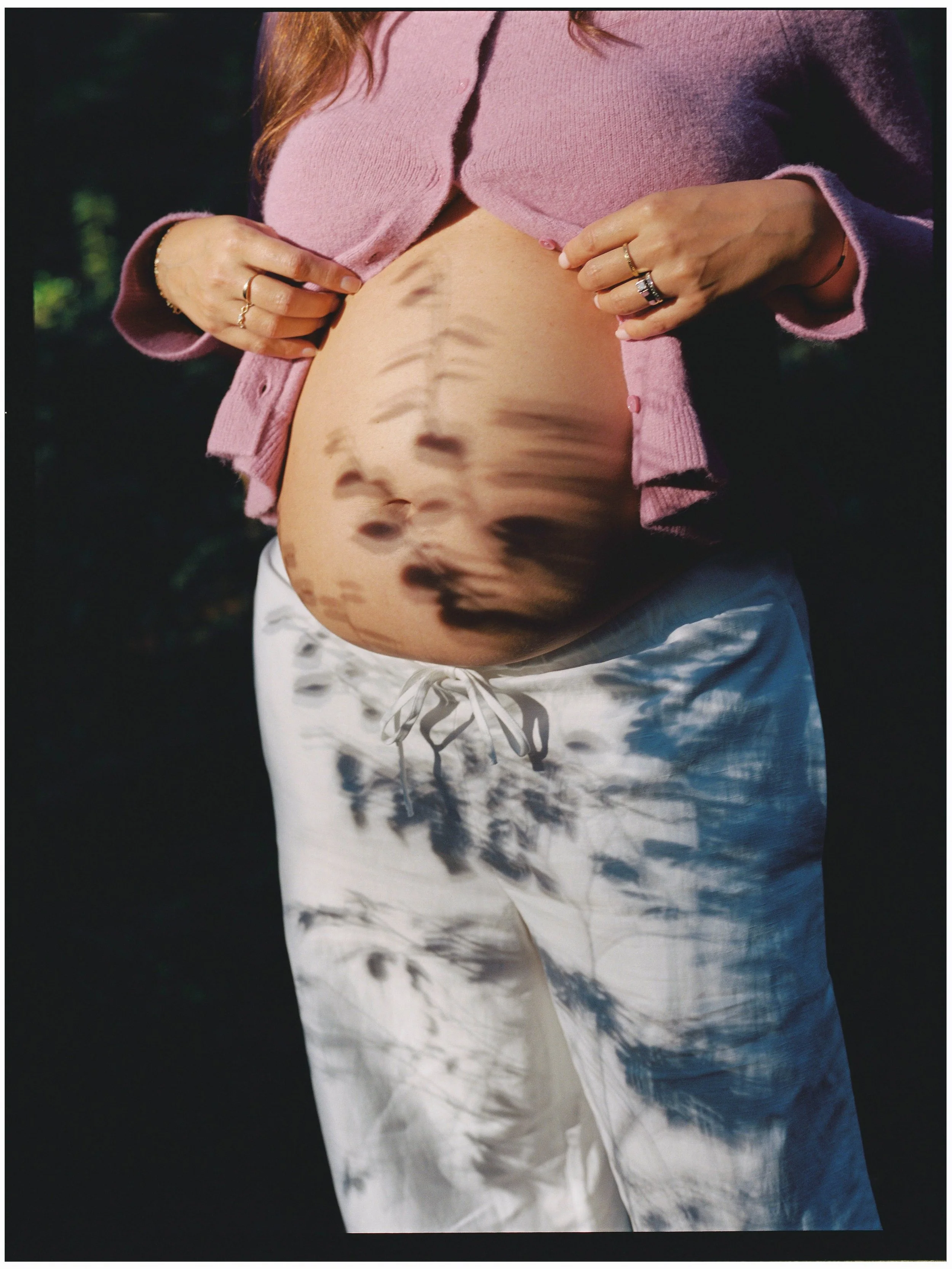 A pregnant woman outdoors flipping up her pink sweater to reveal her belly, which has a shadow reflection of tree branches and leaves.