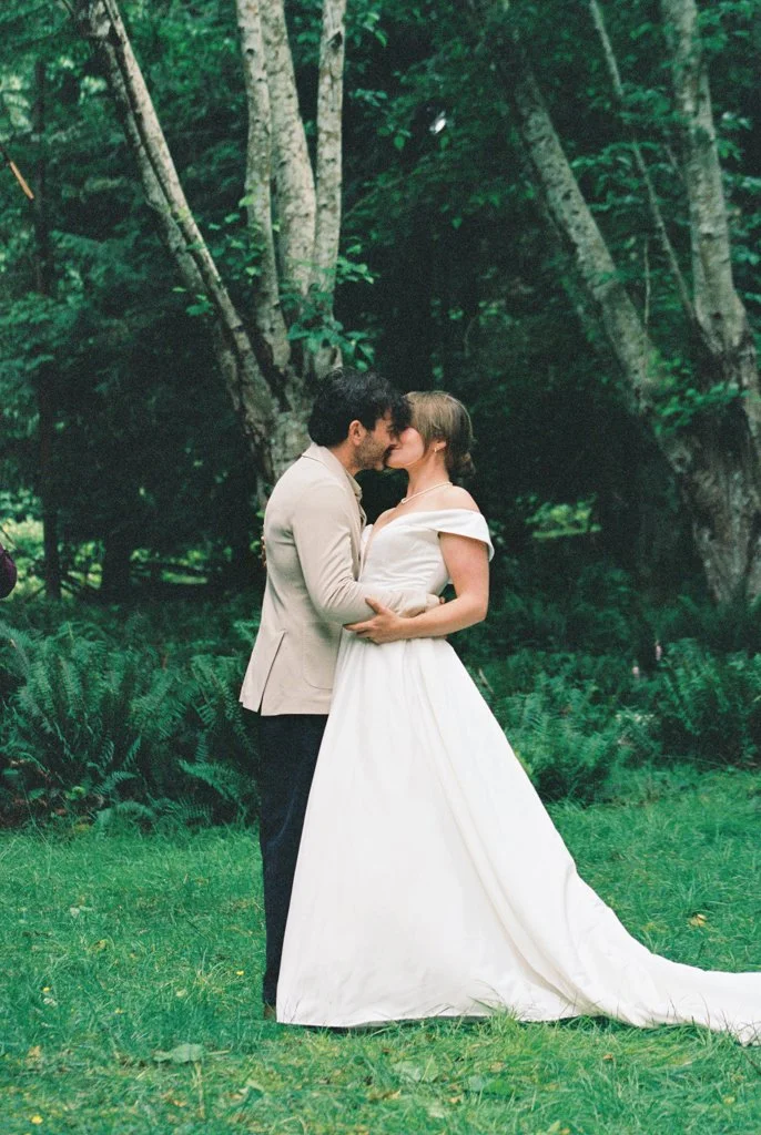 A bride and groom kissing outdoors in a forest setting, with green grass and trees in the background.
