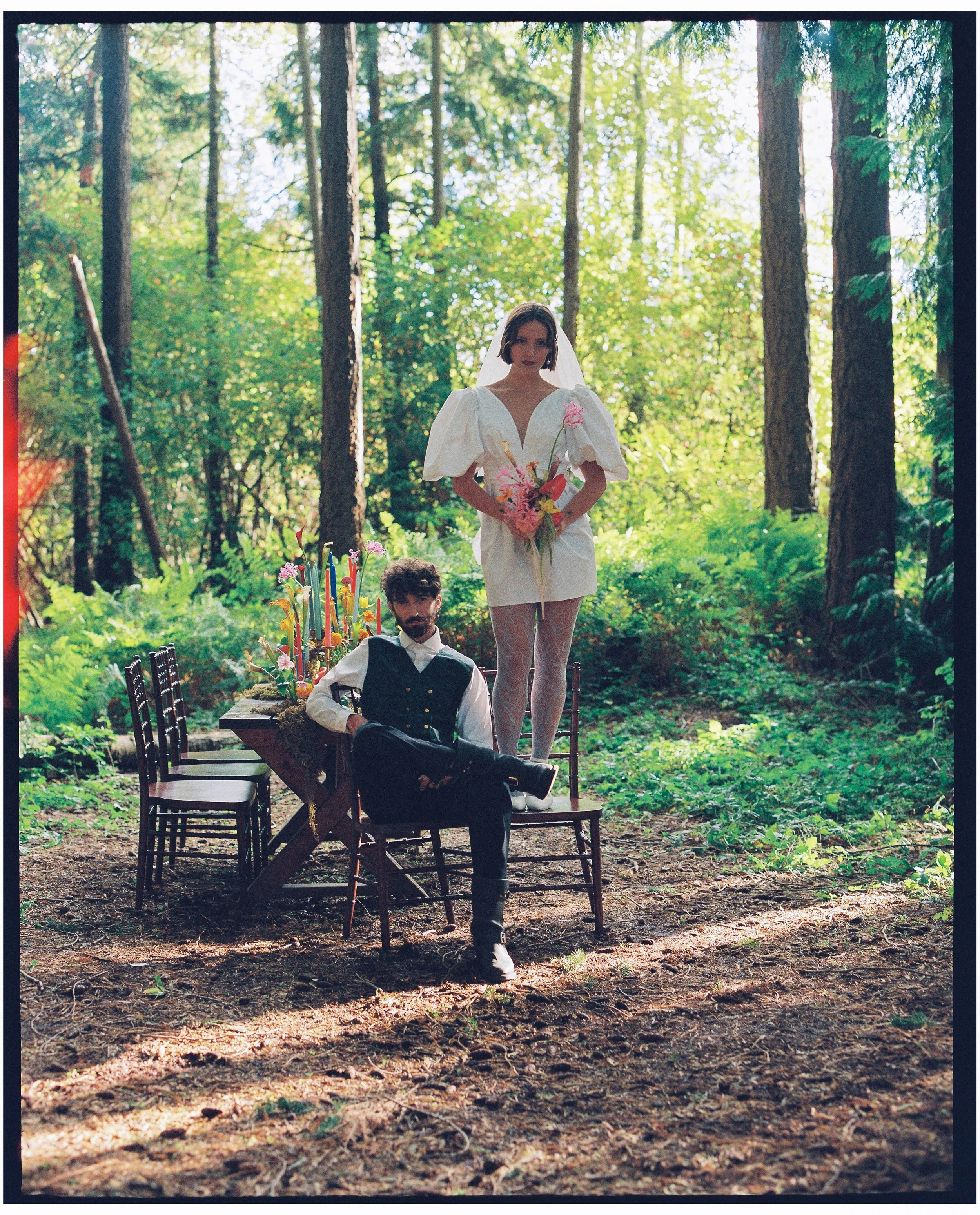 A woman in a white dress and veil holding pink flowers standing on a chair and a man in a black vest and white shirt sitting on a chair in a forest setting with a decorated table with candles and flowers nearby.