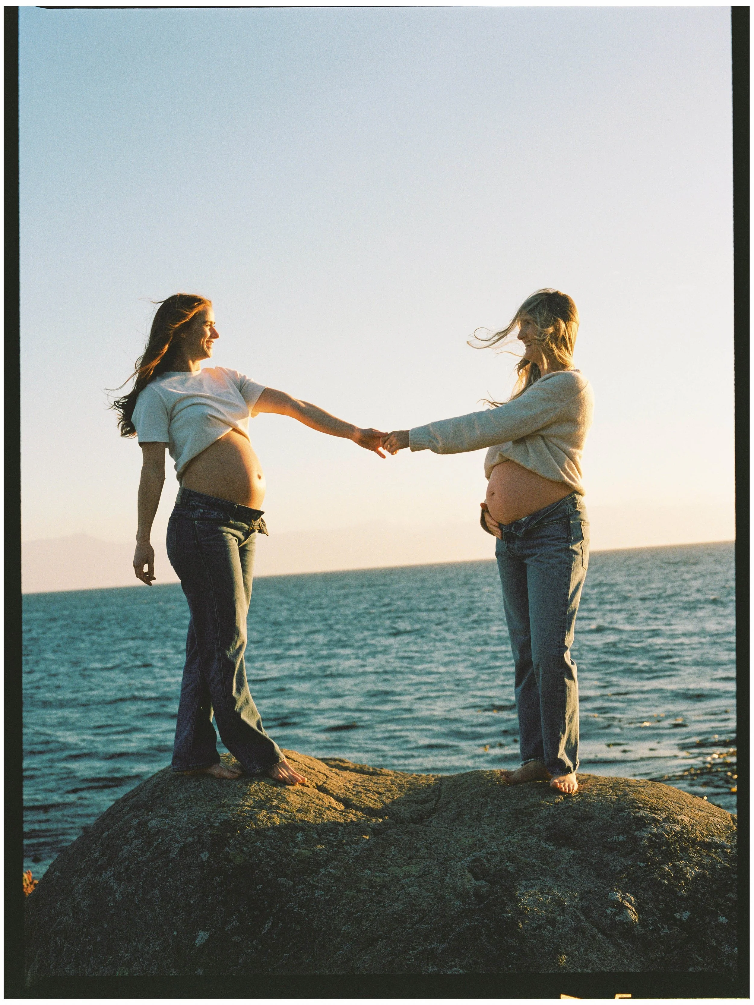 Two pregnant women standing barefoot on a large rock by the ocean, reaching out and holding hands at sunset.