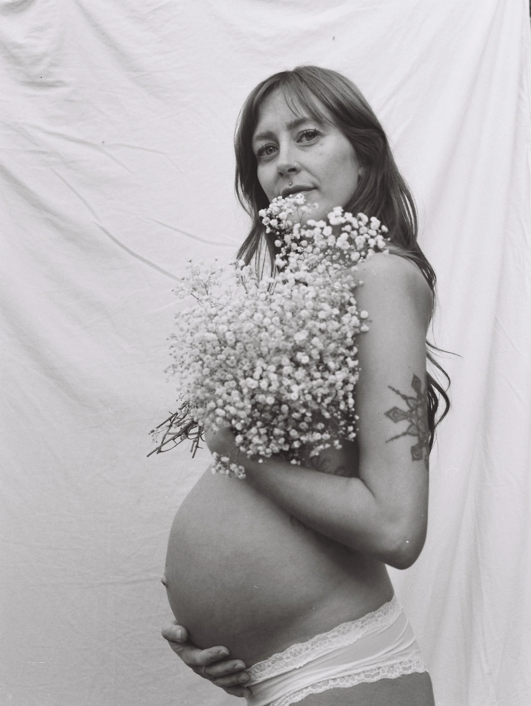 A pregnant woman with long hair holding a bouquet of baby's breath flowers, wearing white lace underwear, standing against a plain white backdrop.