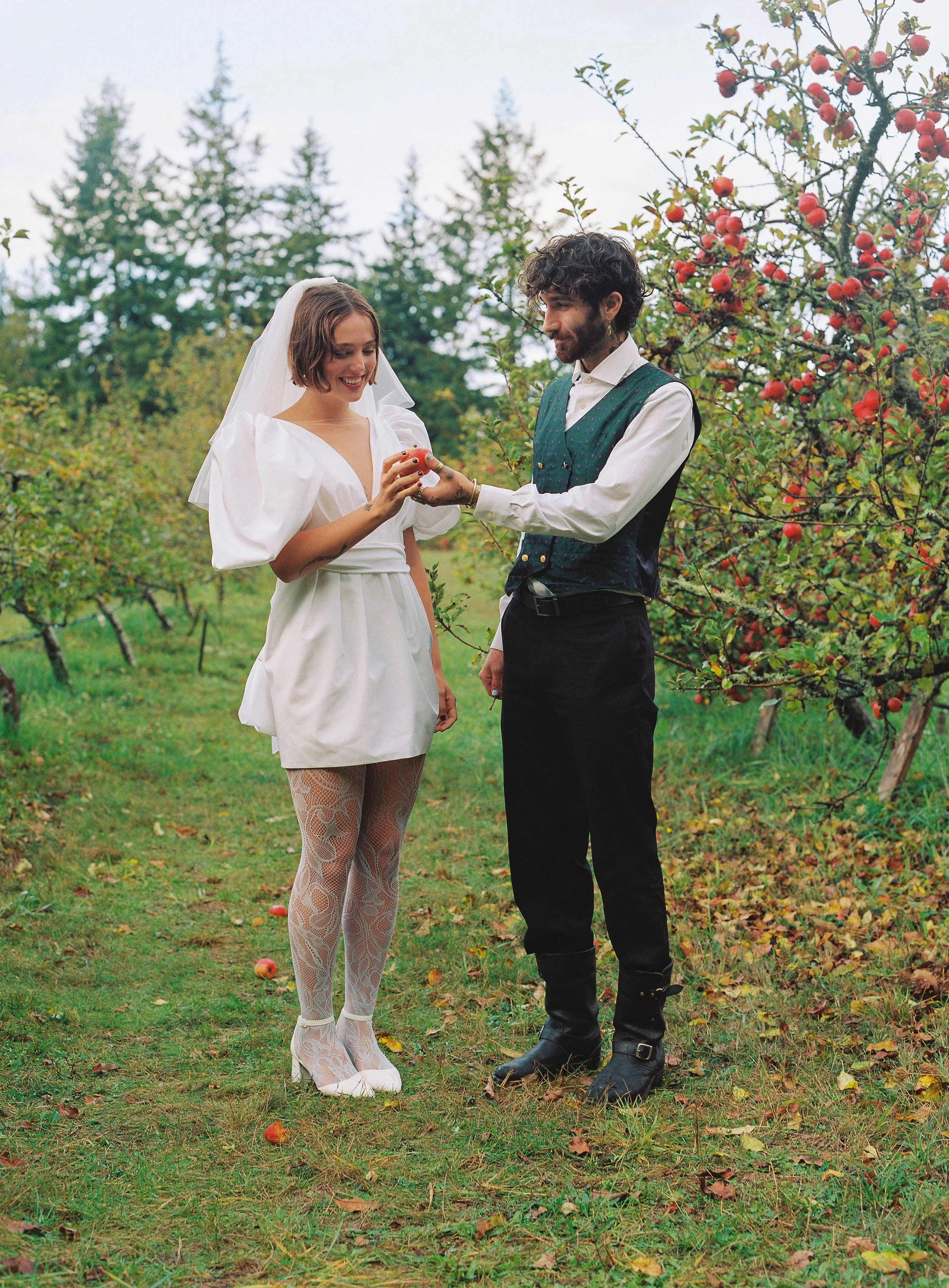 A woman in a white wedding dress and lace tights standing beside a man in vintage clothing, holding apples in an orchard with trees and grass, suggesting a wedding or romantic scene.