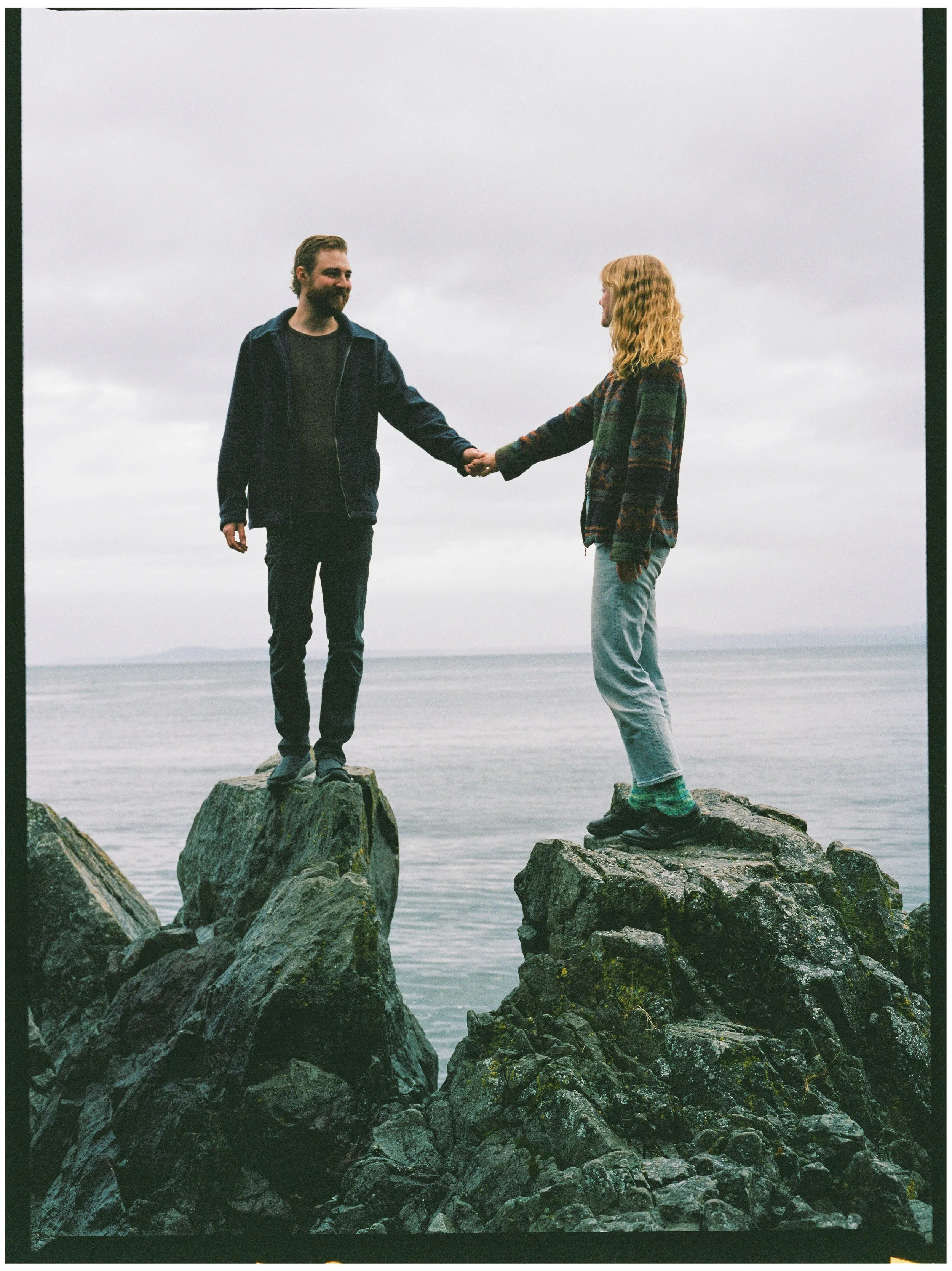 A man and a woman holding hands while standing on separate rocks near a body of water under an overcast sky.