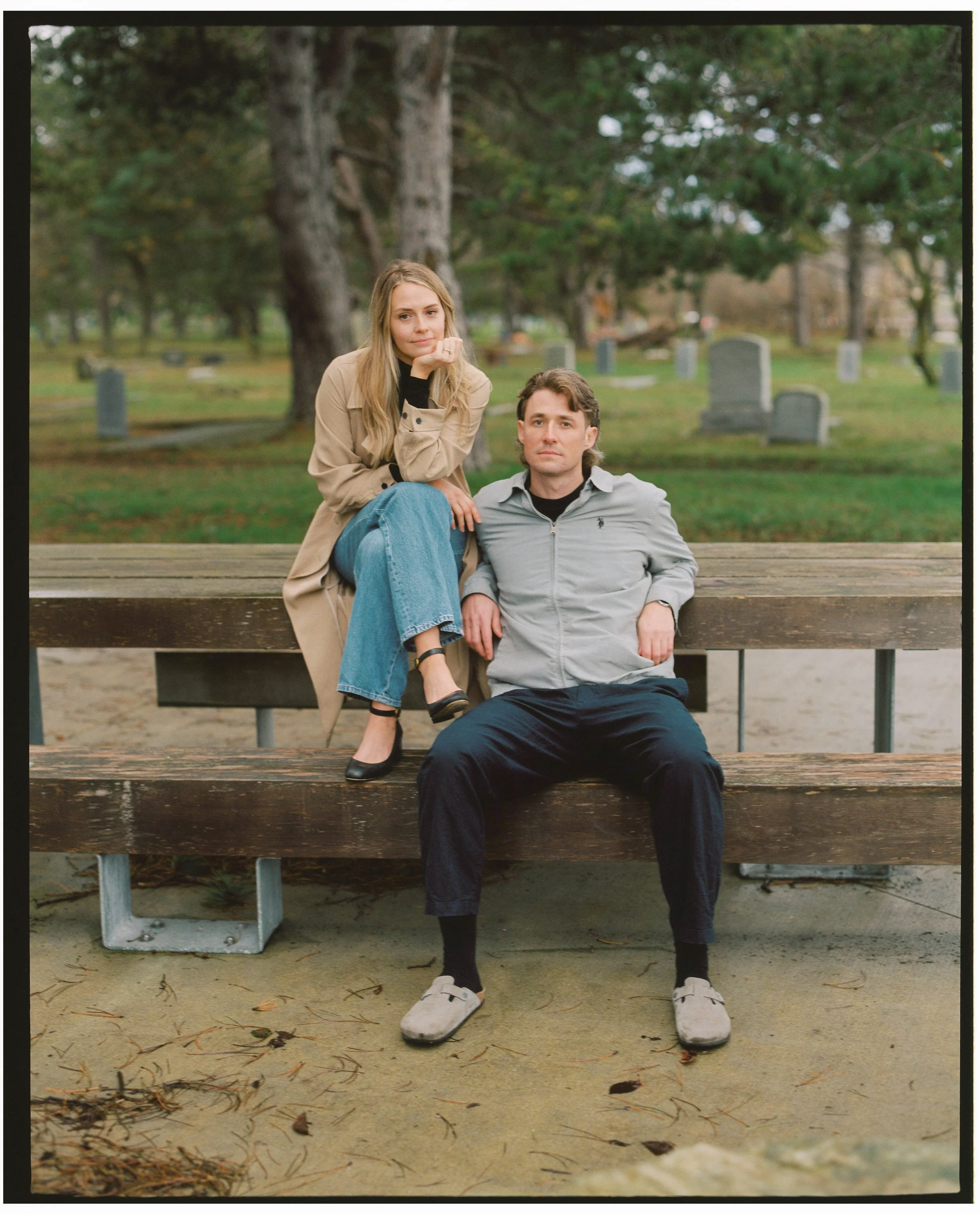 A woman and a man sitting on wooden benches in a park with trees and gravestones in the background. The woman is sitting on the top bench, leaning on her hand, while the man is sitting on the lower bench with his hands resting on his legs.