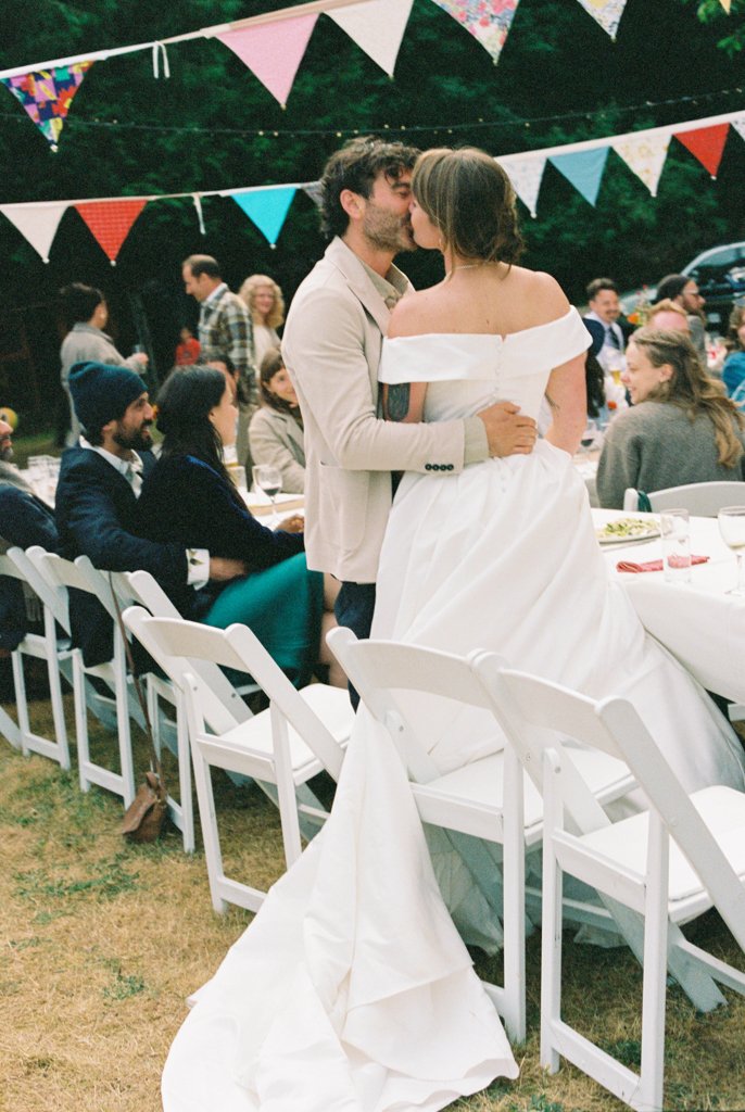 Couple sharing a kiss at a wedding reception, with guests seated at tables under colorful bunting.