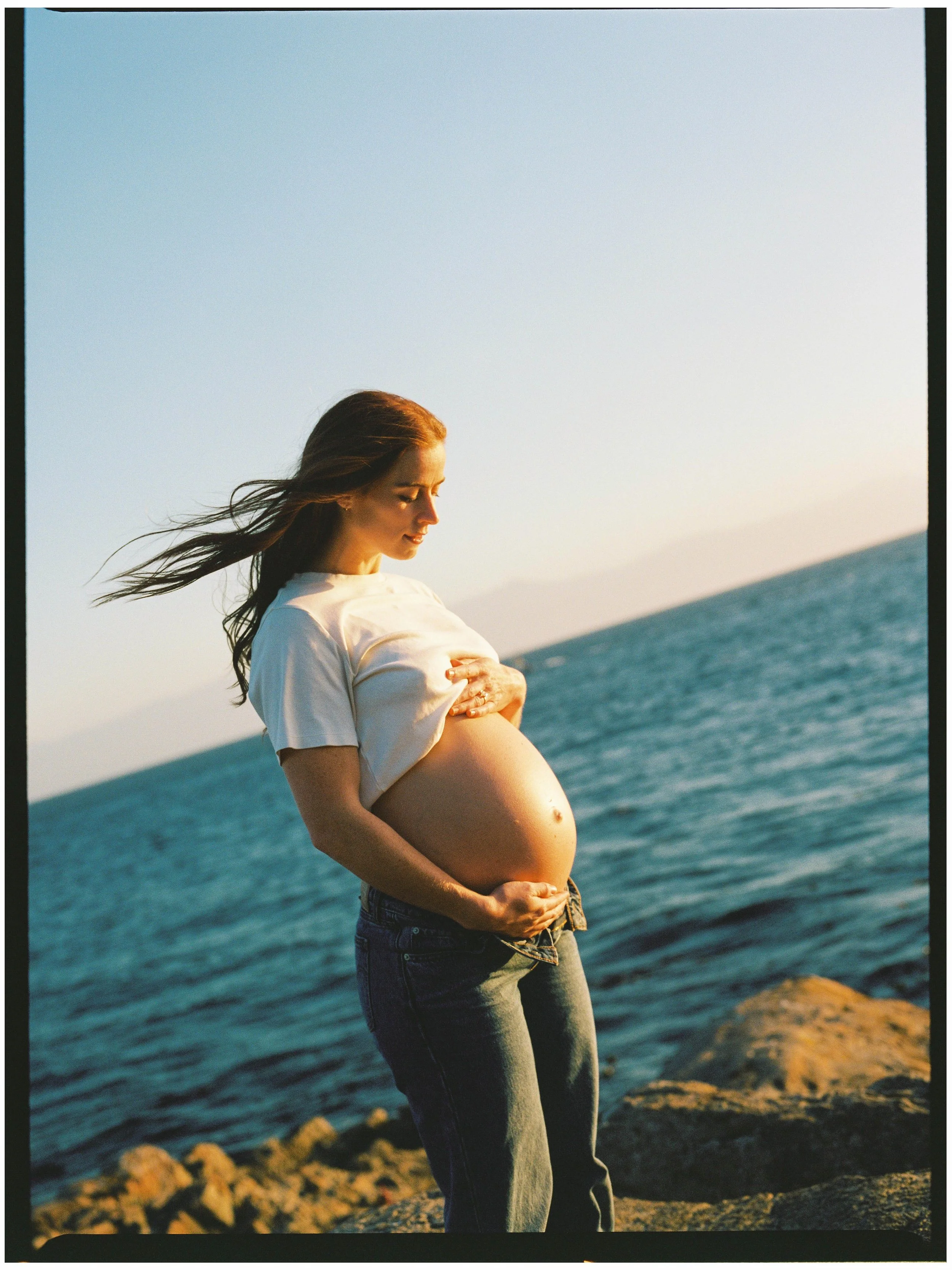 Pregnant woman standing on rocky shoreline at sunset, looking down and touching her belly.
