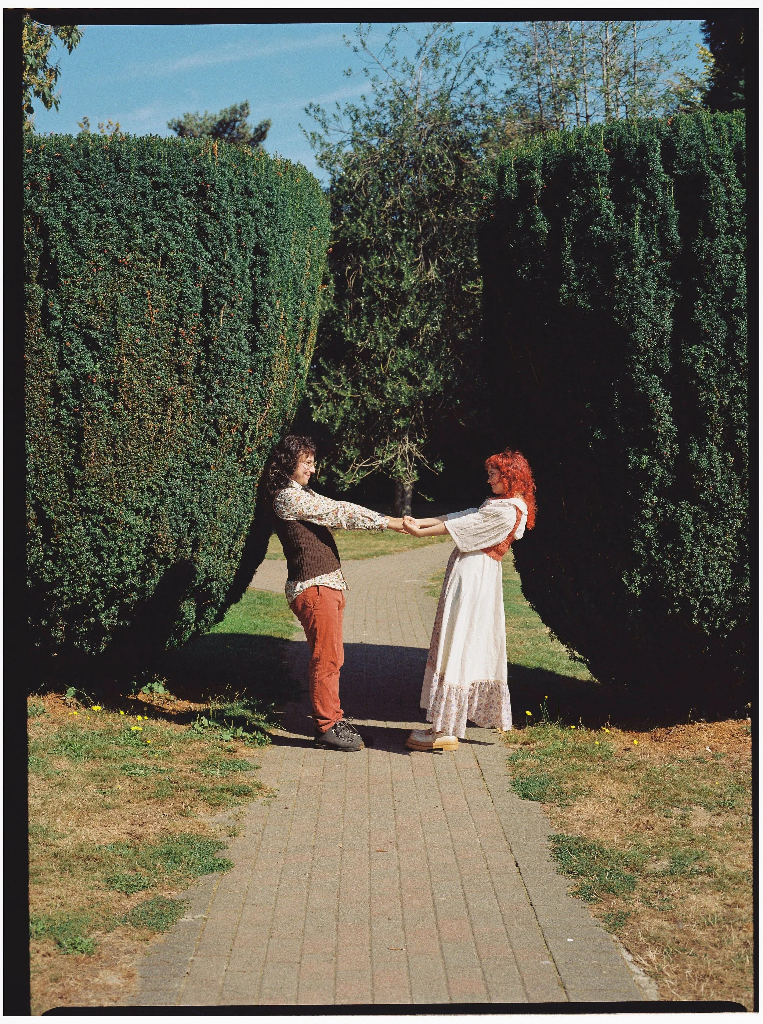 Two women holding hands, facing each other on a brick path, surrounded by large, green, rounded bushes and trees under a clear blue sky.