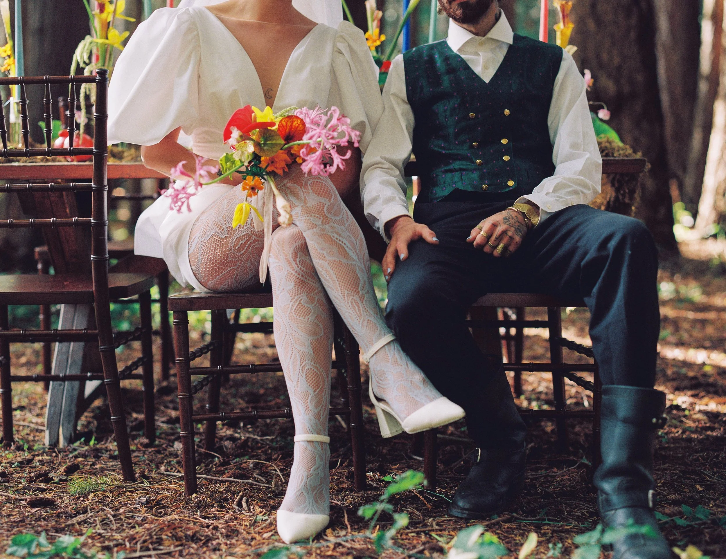 A woman holds a bouquet of colorful flowers while sitting next to a man in a forest setting, both dressed fashionably.