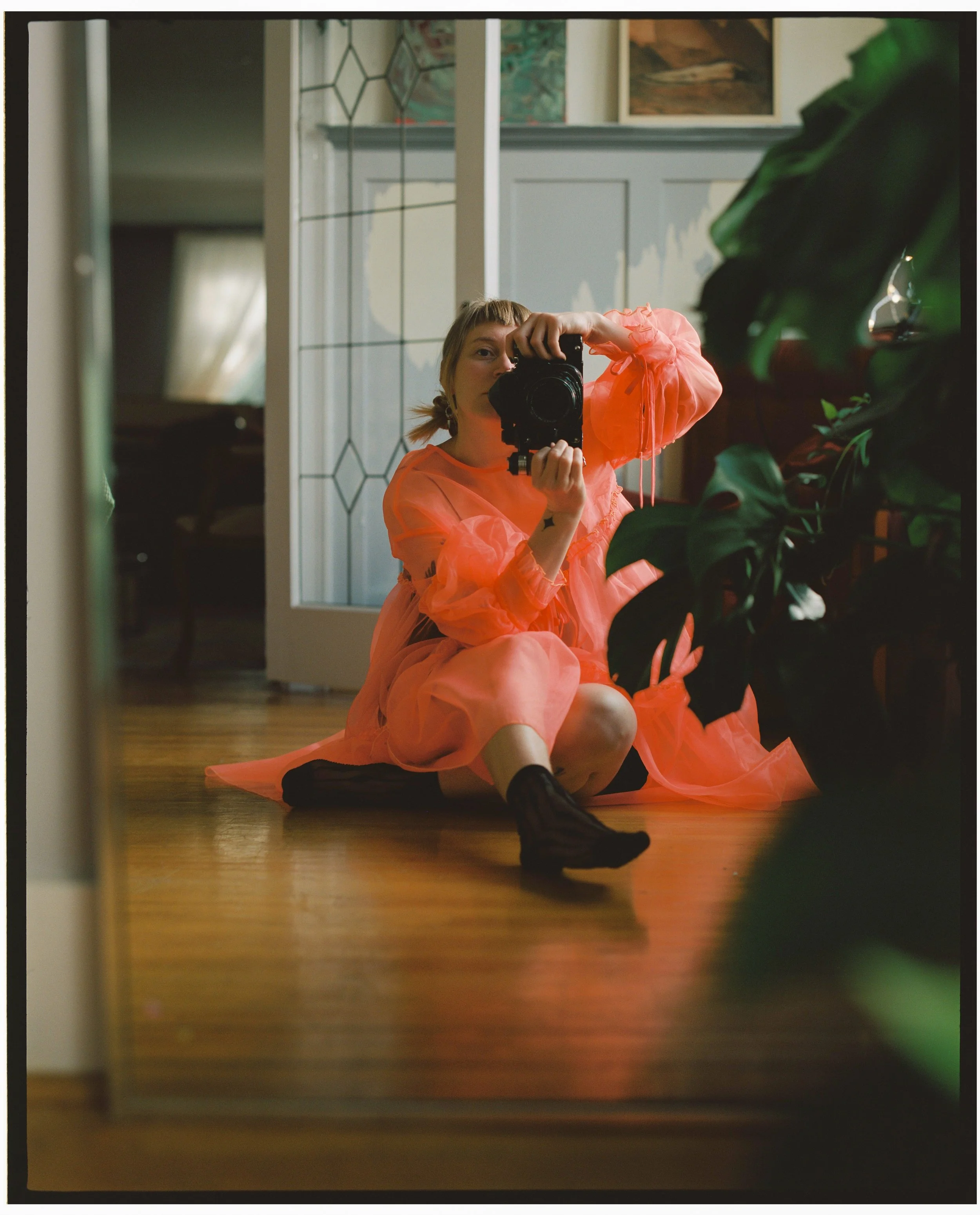 Person sitting on a hardwood floor taking a mirror selfie with a camera, wearing a sheer coral pink dress and black socks, with a green leafy plant partially visible in the foreground.