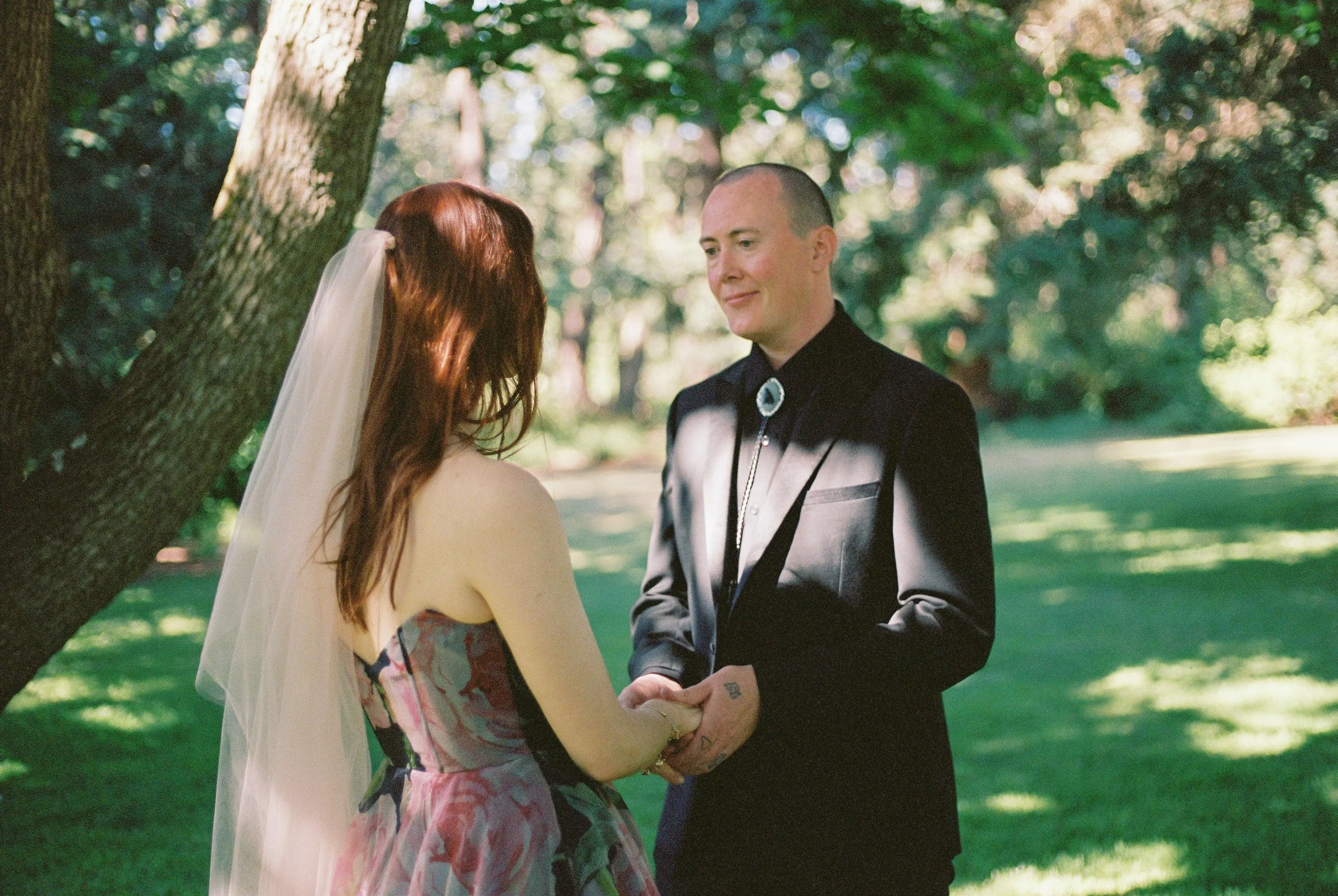 A couple getting married outdoors in a park, holding hands and facing each other, with trees and sunlight in the background.