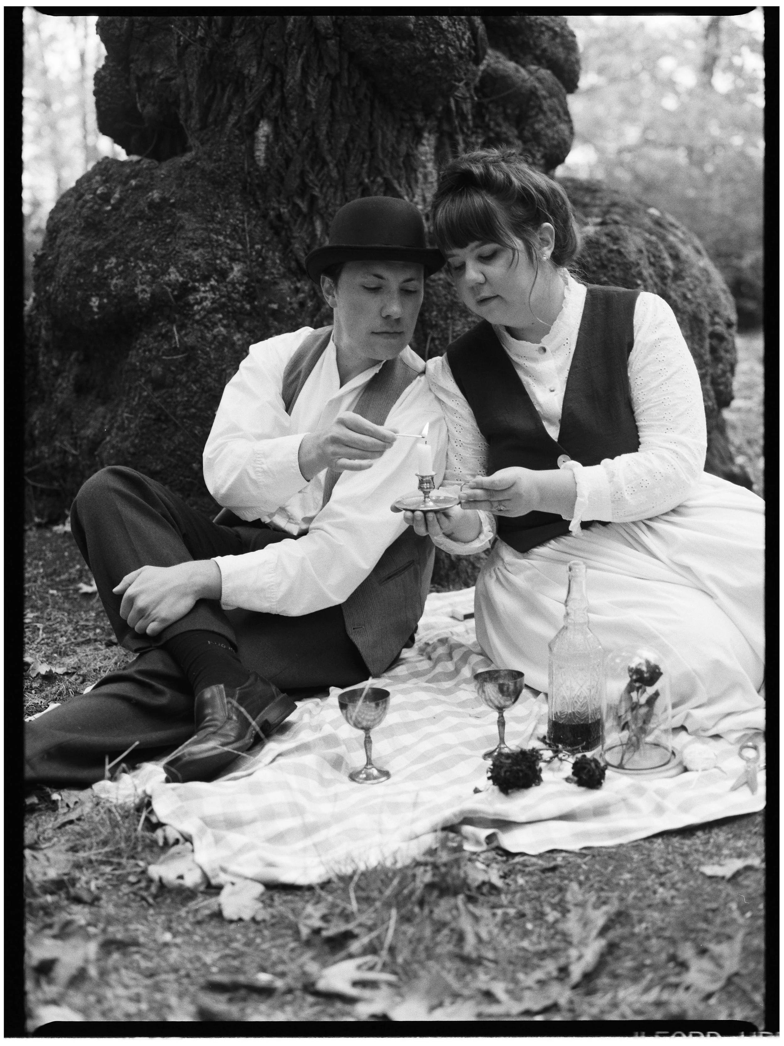 A black-and-white photo of a young man and woman having a picnic outdoors in a wooded area. They are sitting on a checkered blanket, sharing a candle, wine glasses, a bottle, and a small display with flowers. The man is wearing a hat, vest, and dress